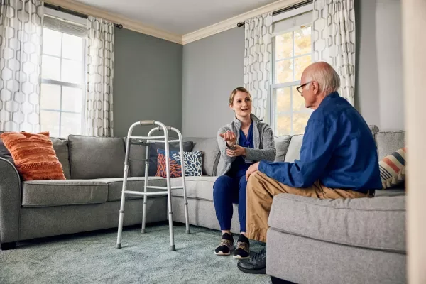 a nurse and older man sitting and talking on a gray sofa in a living room discussing in home care.