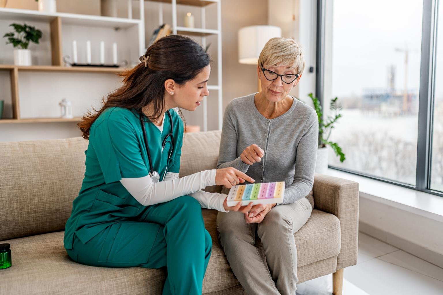 A healthcare professional in medical scrubs consulting with an elderly woman about medication, sitting on a beige sofa in a bright room near a large window.