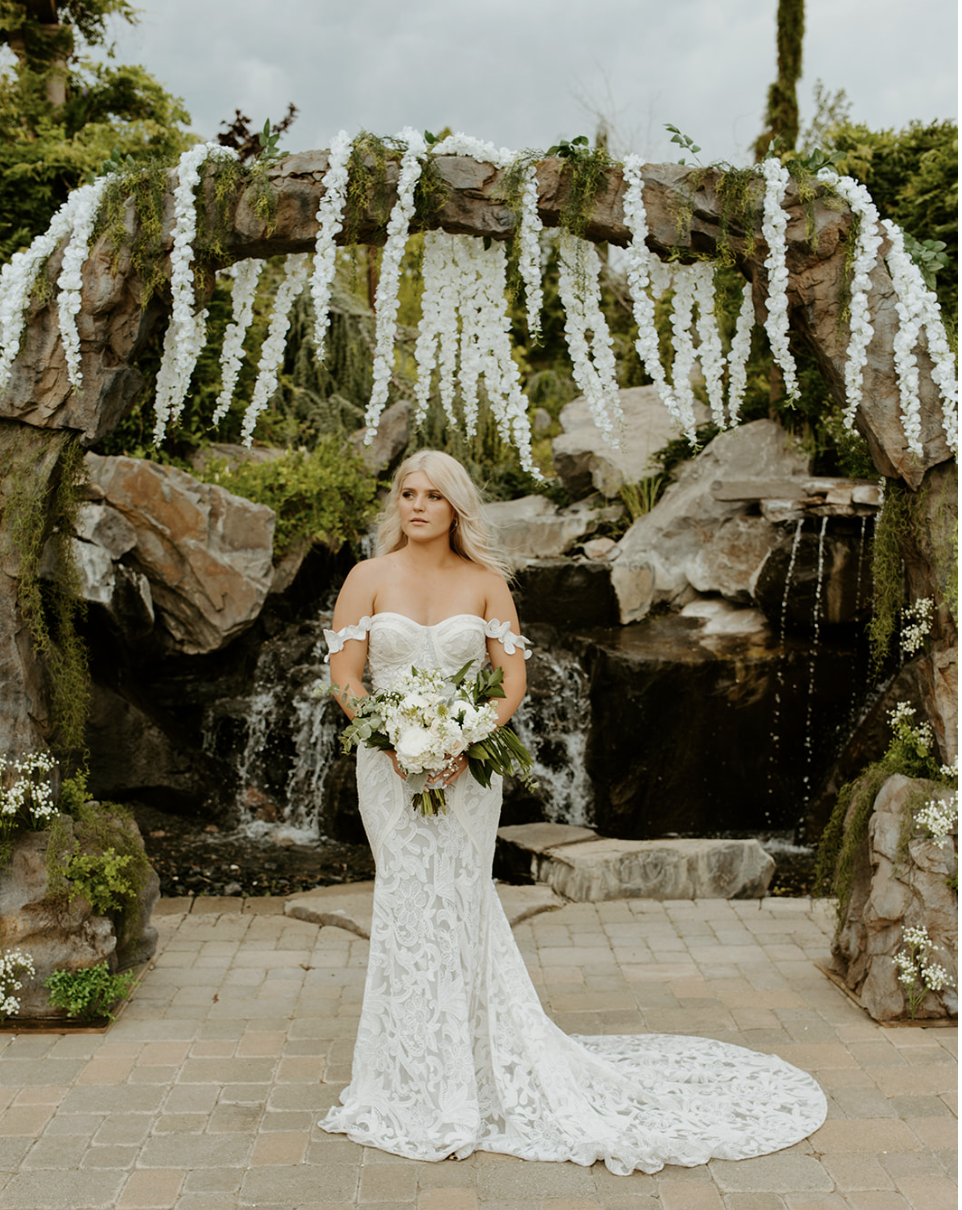 A bride in a white lace wedding gown holding a bouquet of white flowers, standing outdoors beneath a floral arch with white hanging flowers and greenery, with a small waterfall in the background.