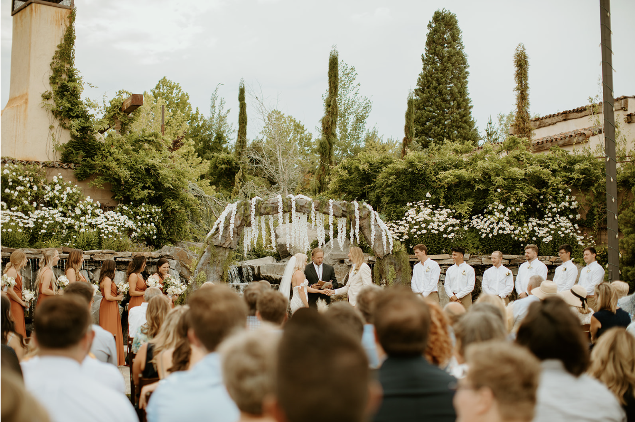 Outdoor wedding ceremony with bride and groom exchanging vows beneath floral arch, surrounded by bridesmaids in rust-colored dresses and groomsmen in white shirts, with guests seated in front, set in a garden with trees and white blooming flowers.