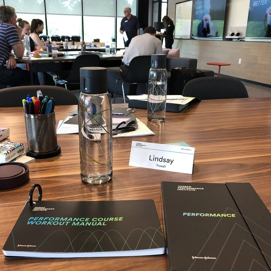 A conference room with a wooden table, notebooks, water bottles, pens, and a nameplate that reads 'Lindsay Troxell'. There are people seated at the table and others standing or walking in the background, with large windows and digital screens on the 