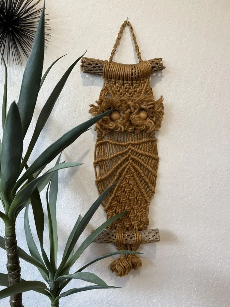 Decorative wall hanging made of woven and knotted rope, resembling a gnome with a round nose, eyes, a beard, and a pointed hat, mounted on a textured white wall connected to two small wood pieces at the top and bottom.