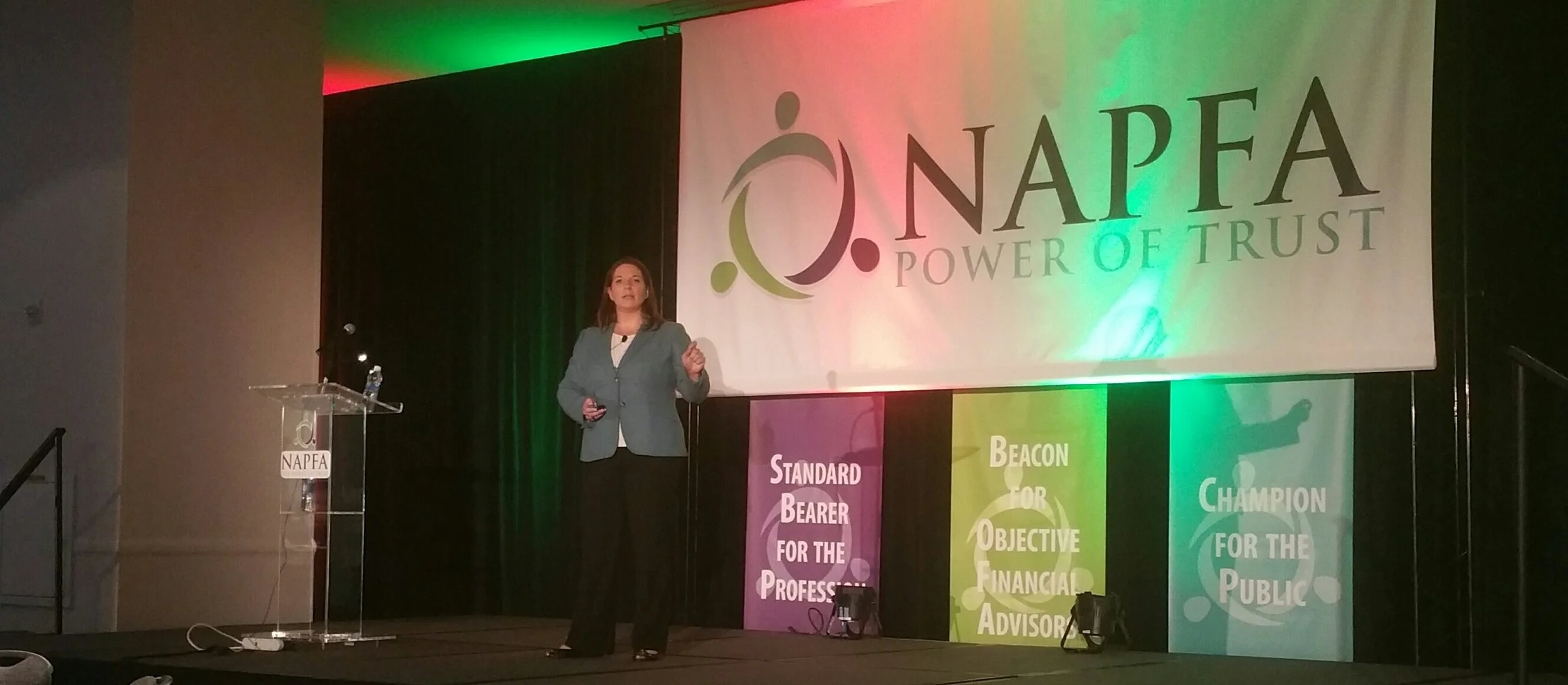 A woman is giving a presentation on stage at a NAPFA event, with a large screen behind her displaying the NAPFA logo and the tagline 'Power of Trust'. There are colorful banners on the stage with words like 'Standard Bearer for the Profession', 'Beac