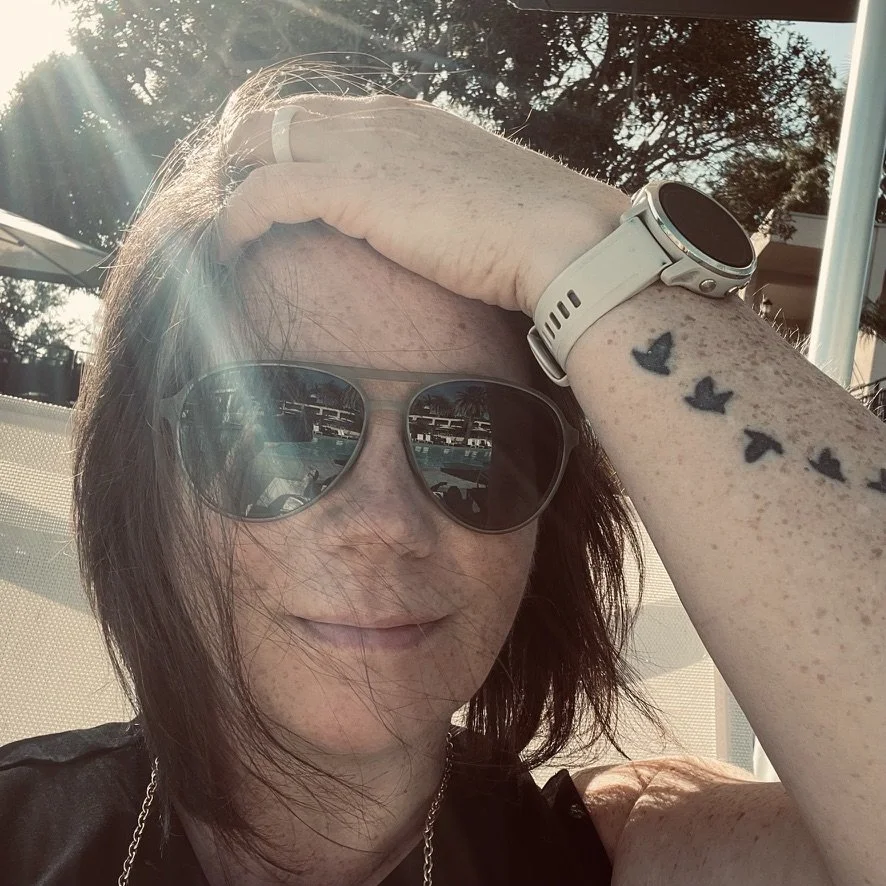 Lindsay Troxell with dark hair wearing sunglasses, a white wristwatch, and a black top, smiling with her hand on her forehead, sitting outdoors near a pool with trees in the background, sunlight shining.
