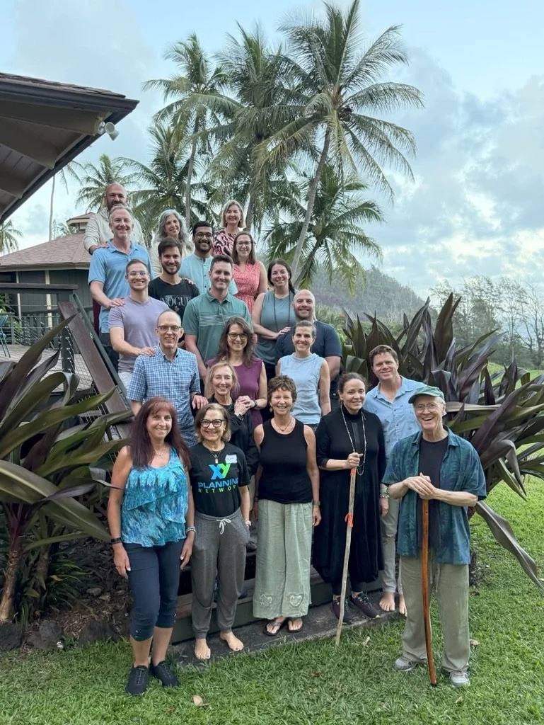 Group of 20 people gathered outdoors on a stone staircase surrounded by tropical plants and tall palm trees, with cloudy sky overhead.