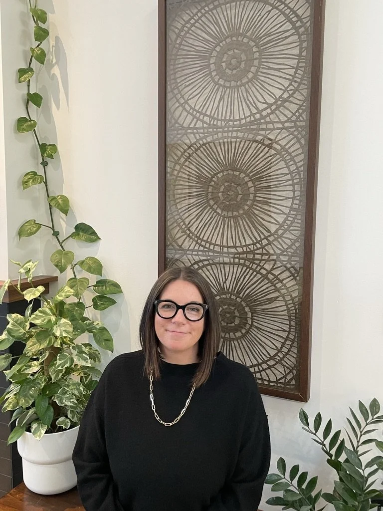 Lindsay Troxell with shoulder-length brown hair, wearing glasses, a black sweater, and a gold chain necklace, standing indoors in front of a decorative metal wall art piece and surrounded by large green potted plants.