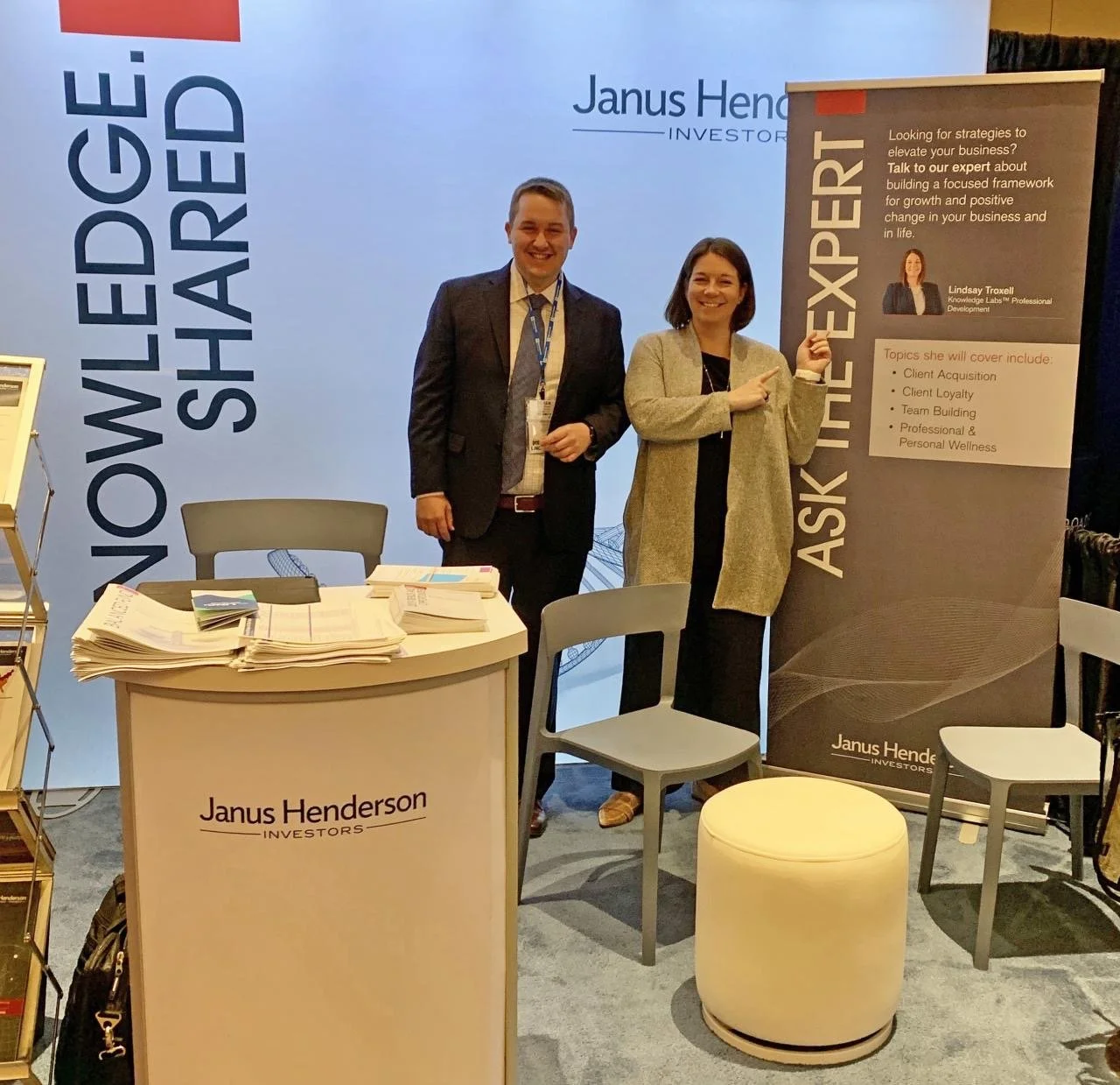 Two people, a man in a suit and a woman in a beige coat, standing at an exhibition booth for Janus Henderson Investors. The woman is pointing to a black roll-up banner that provides details about strategic business growth and professional development