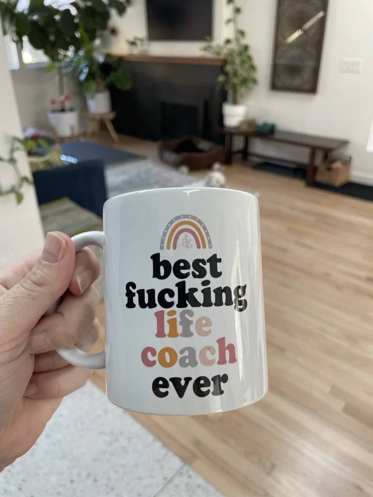 A white mug with colorful text reading 'best f***ing life coach ever' being held in a hand inside a living room with wooden floors, plants, a couch, and a dog bed in the background.
