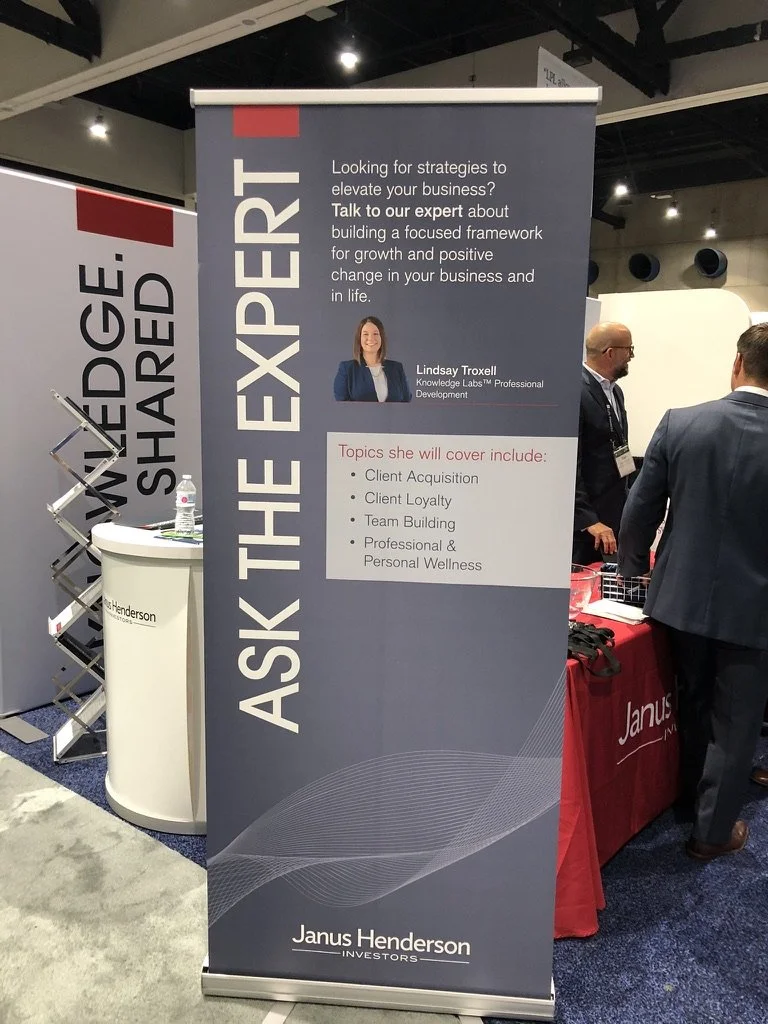 A vertical banner at a business conference promoting Lindsay Troxell, a professional at Knowledge Labs, offering strategies to elevate business, including topics like client acquisition, client loyalty, team building, and wellness. The banner is blue