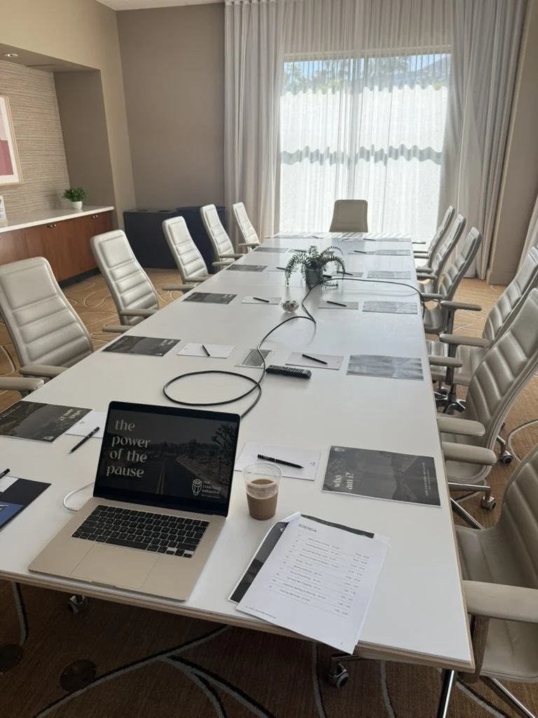 Empty conference room with a long white table, beige chairs, a laptop, documents, a remote, a cup of coffee, and a plant in the center; large window with sheer curtains.