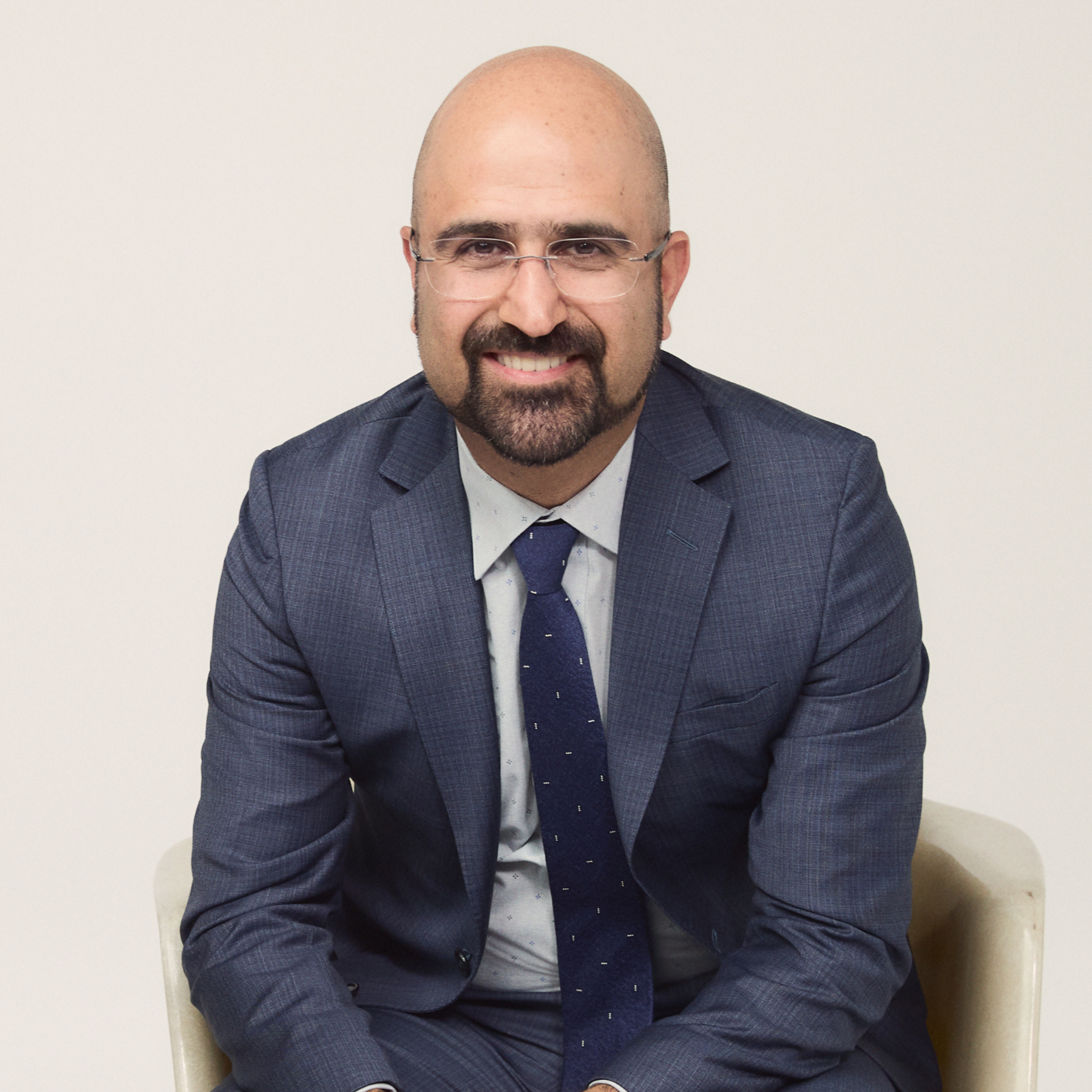 A man with glasses, a beard, and a mustache wearing a blue suit and tie, sitting against a plain white background.