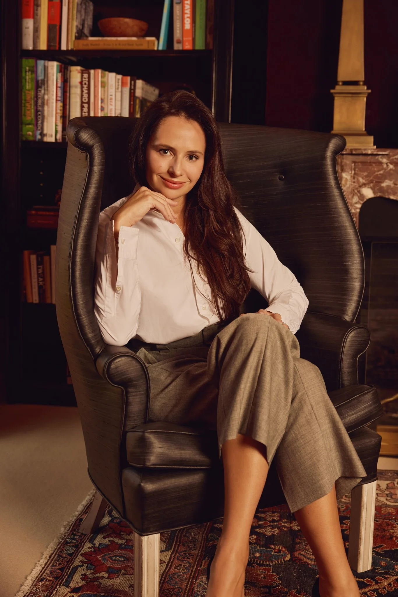 A woman with long brown hair sitting in a black high-back armchair, wearing a white blouse and tan plaid trousers, in a room with bookshelves and warm lighting.