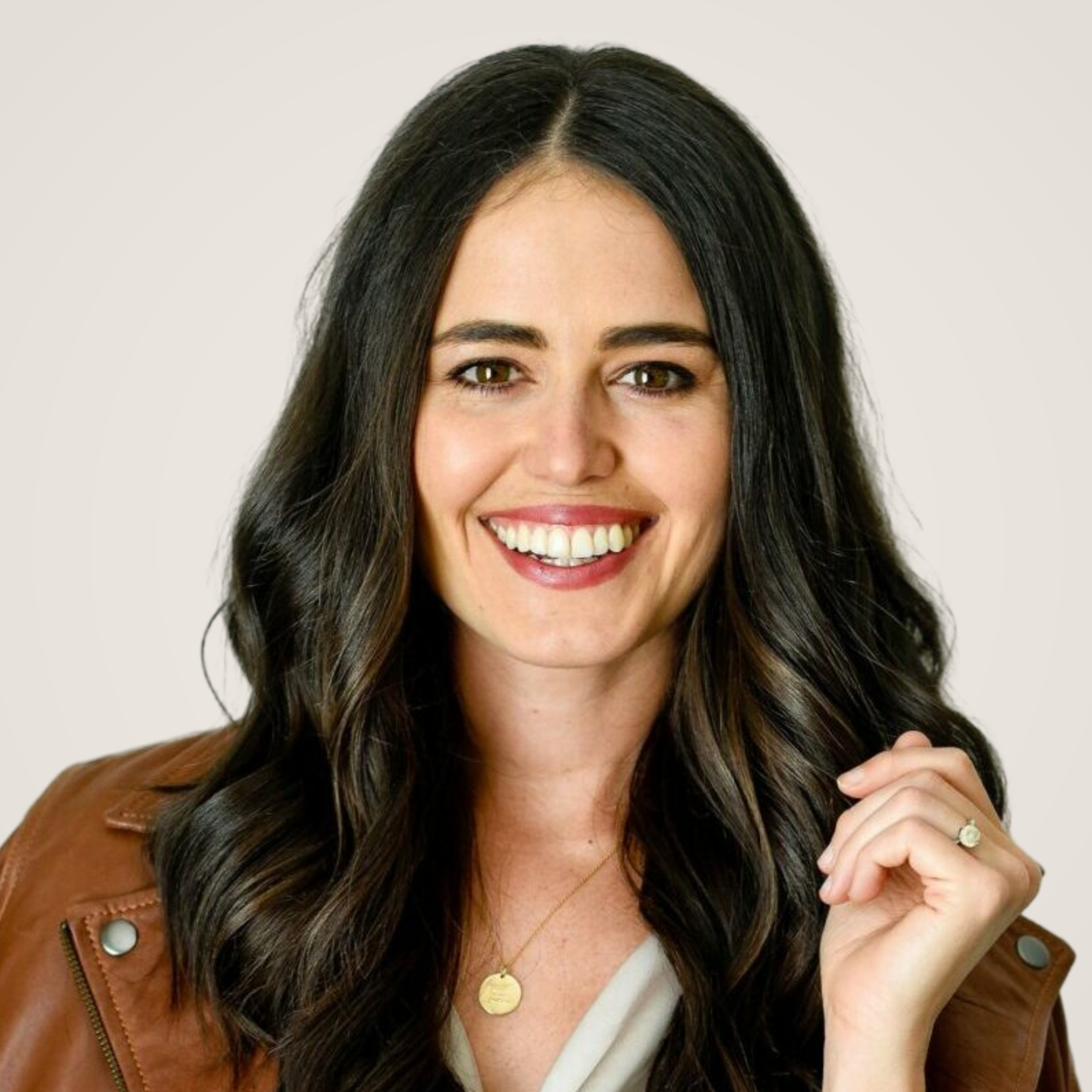 Portrait of a smiling woman with long dark hair, wearing a brown leather jacket, gold jewelry, and a white top, on a plain light background.