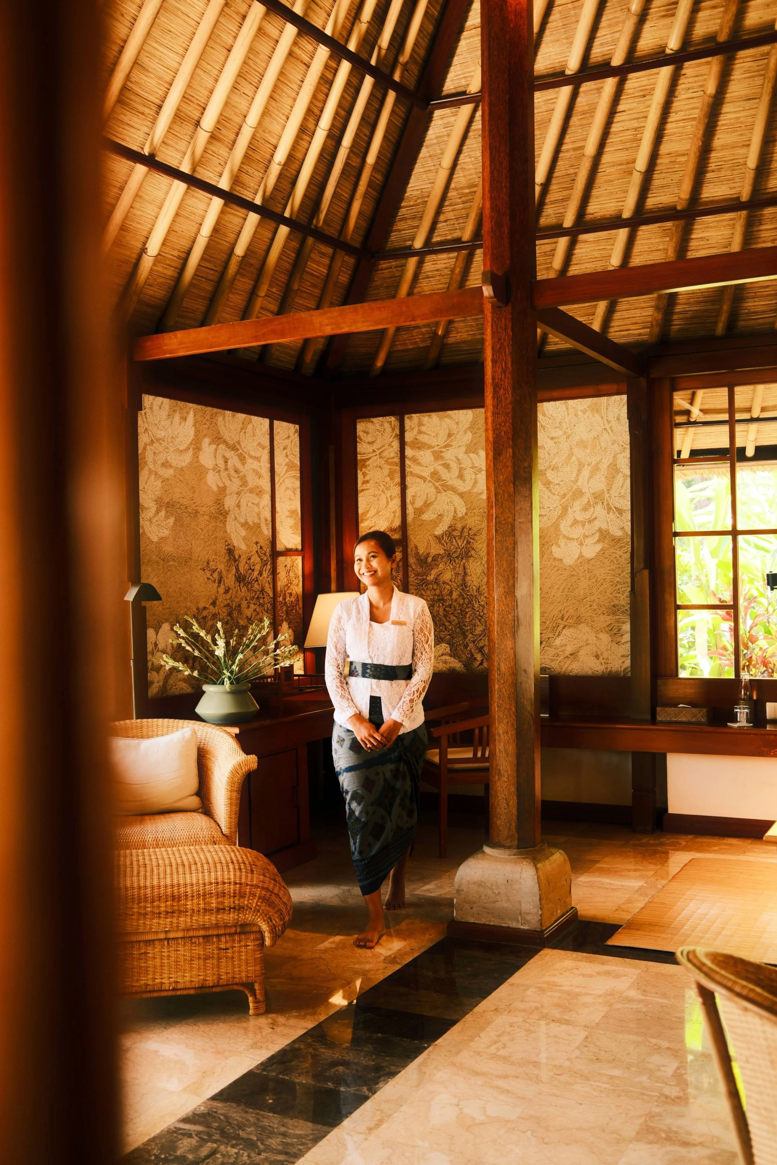 A woman dressed in traditional Balinese attire in a suite at Amandari in Ubud, Bali, standing barefoot in a tropical-themed room with wooden beams, floral wallpaper, and natural sunlight streaming through windows.