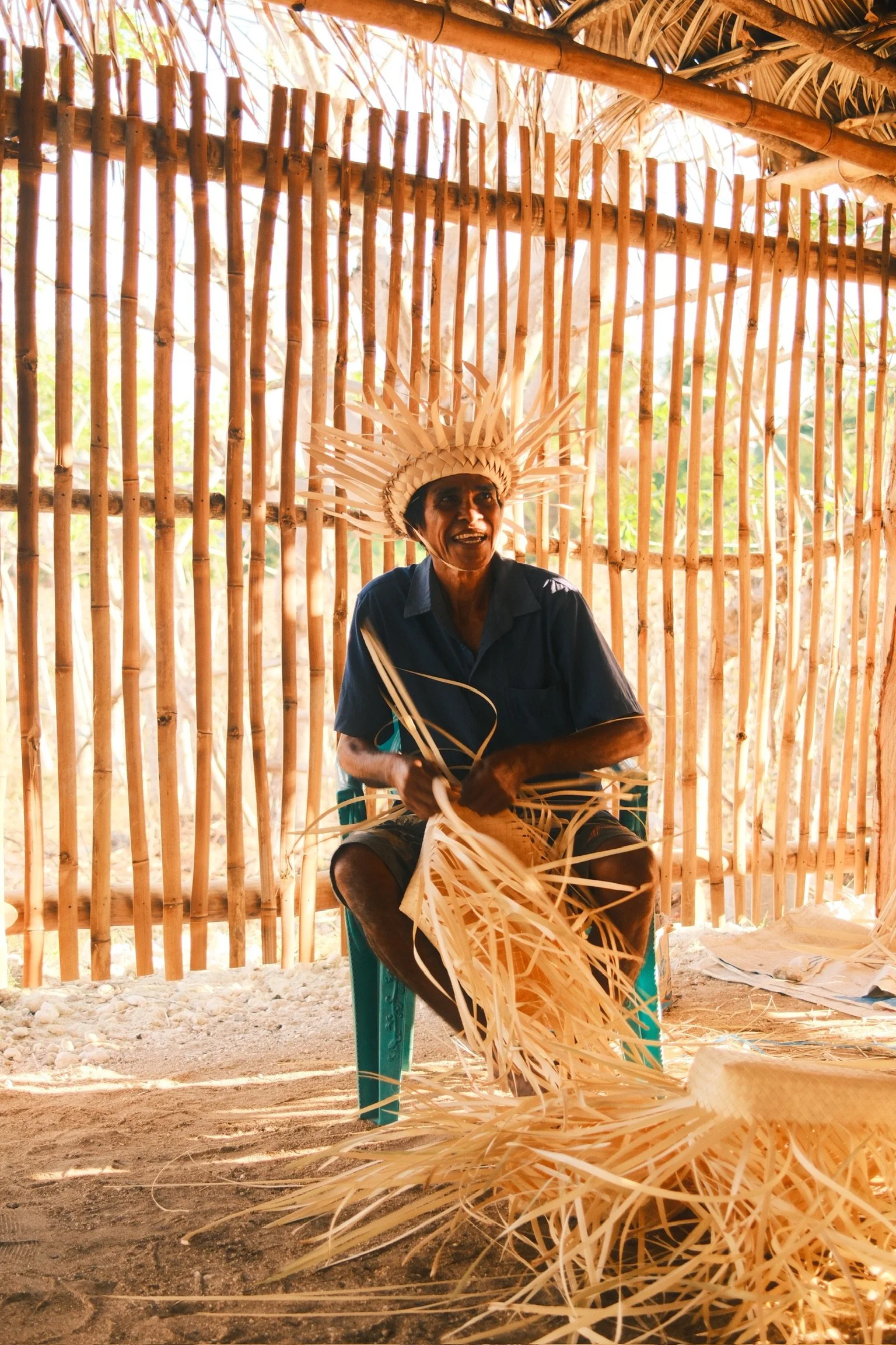 A smiling Rotenese craftsperson weaving palm leaves while wearing a handcrafted headdress.