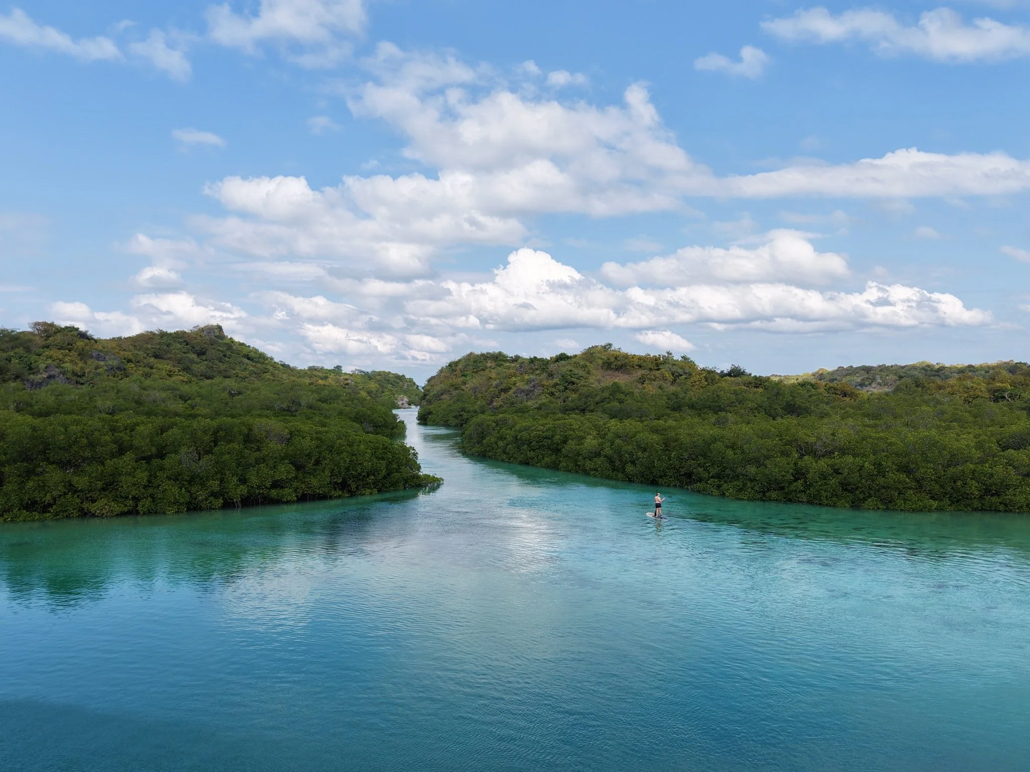 Matt paddle boarding  crossing a calm turquoise channel surrounded by dense mangroves.