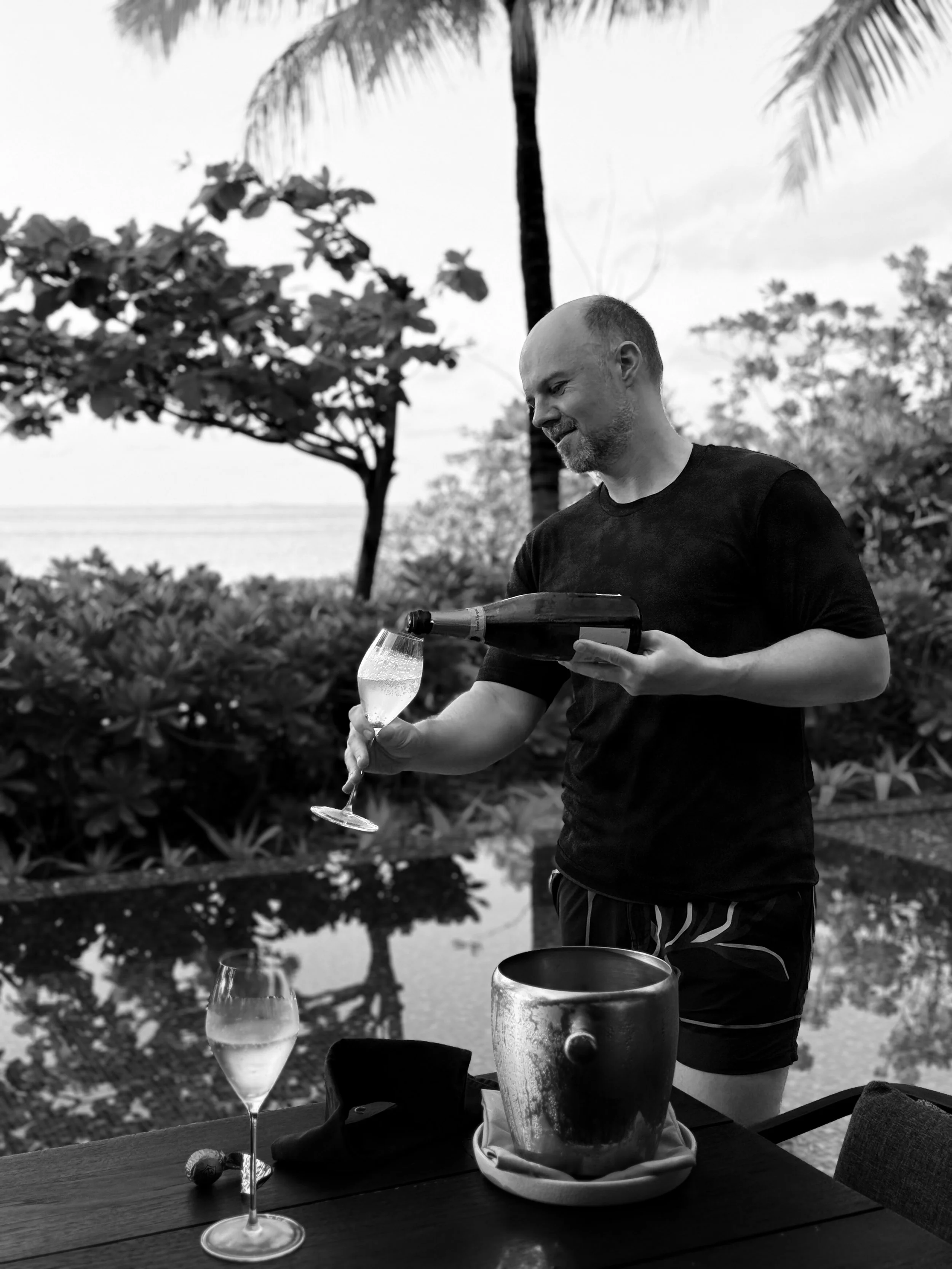 A man with a shaved head and beard, wearing a black t-shirt, is pouring a drink from a bottle into a wine glass outdoors near water and surrounded by trees and palm leaves.