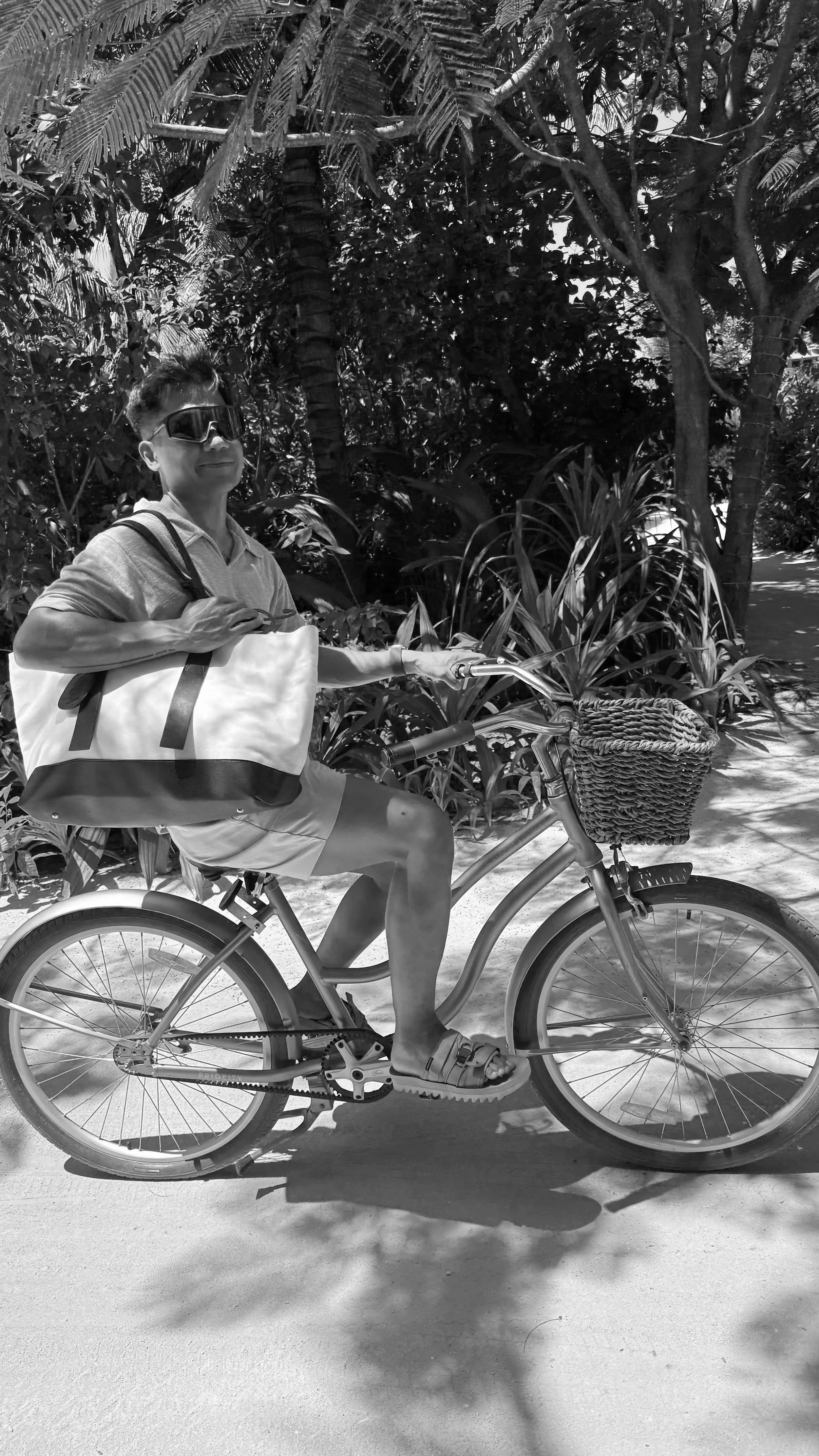 A person riding a bicycle on a path surrounded by lush tropical plants and trees. The rider is wearing sunglasses, a polo shirt, shorts, and sandals, and has a large tote bag over their shoulder. The bicycle has a front basket, and the photo is in black and white.