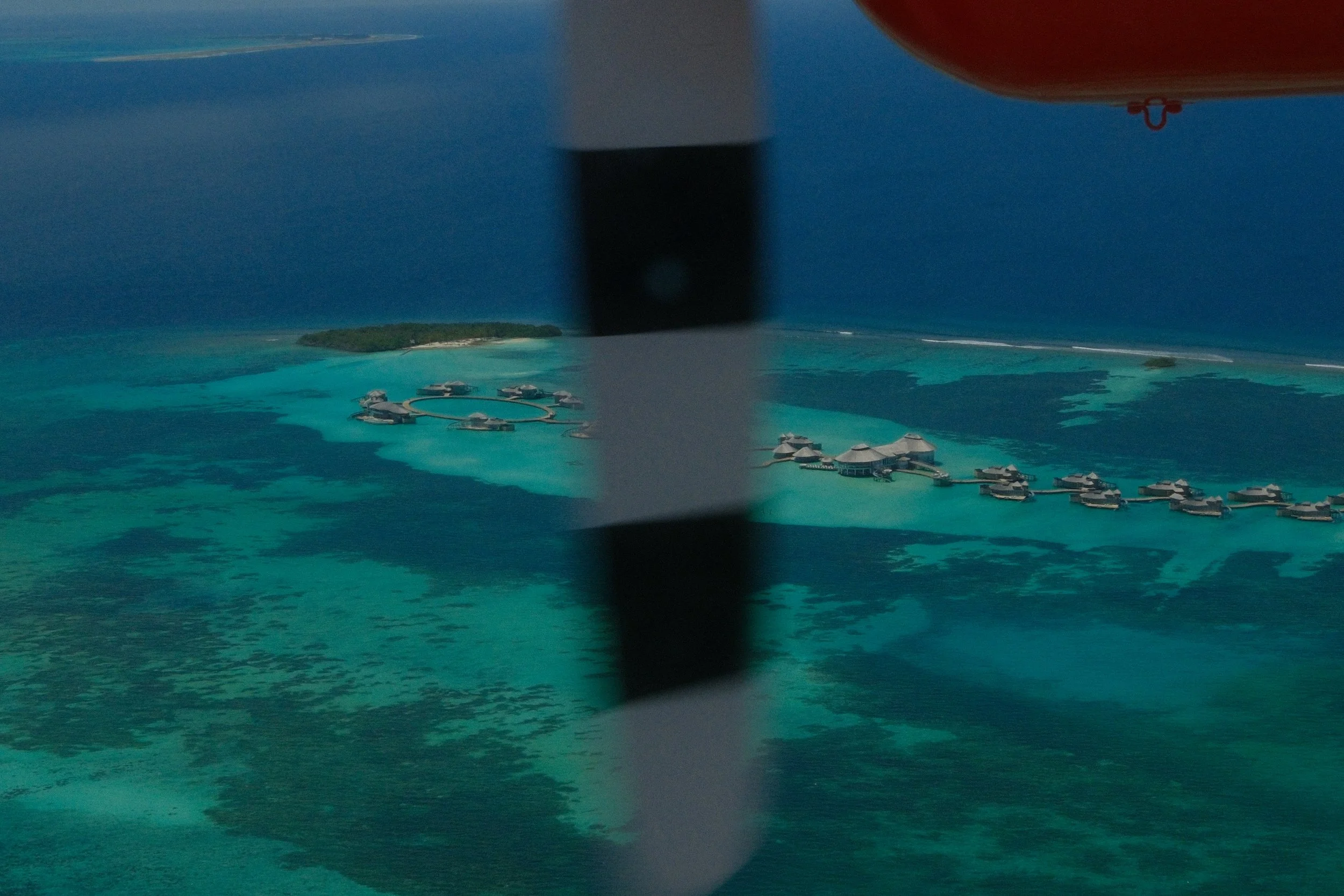 Aerial view of turquoise tropical lagoon with overwater villas and a blurred seaplane propeller in the foreground.