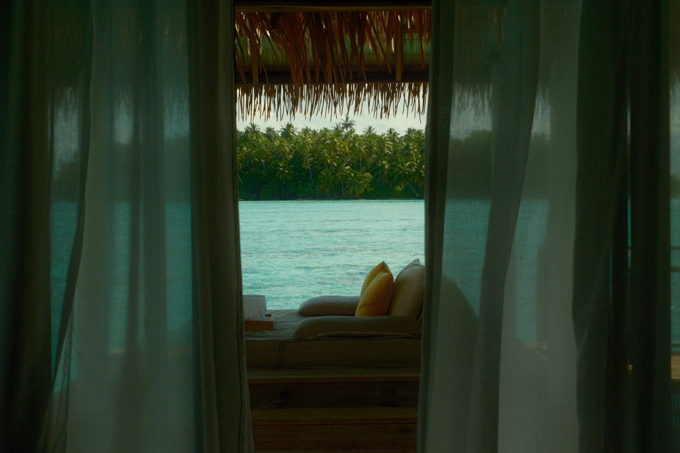 A cushioned daybed overlooking the turquoise lagoon and a palm-covered island through sheer curtains.