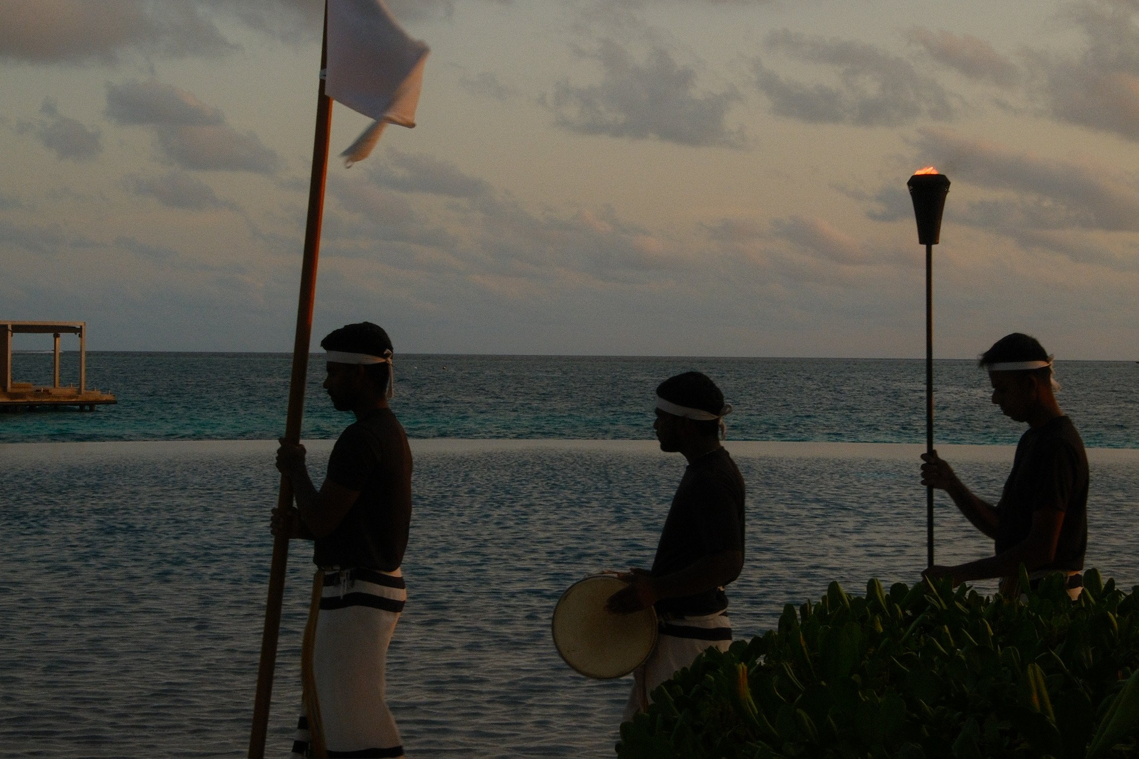 Ritz-Carlton Maldives ceremony by the ocean at sunset, with musicians and torch bearers silhouetted beside the water.