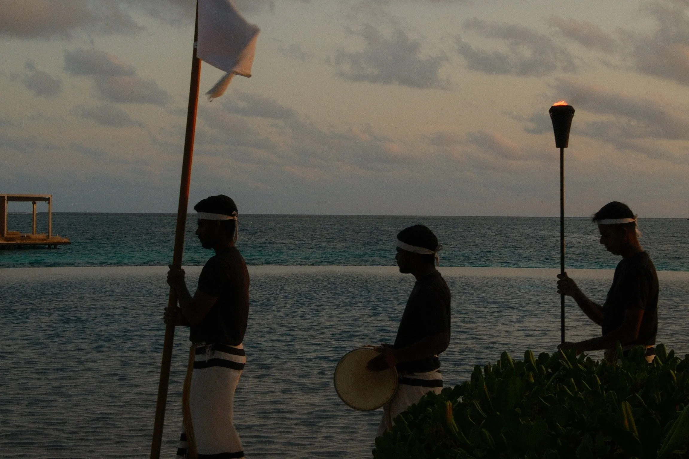 Three resort hosts carrying a flag, drum and flaming torch beside the lagoon at sunset.