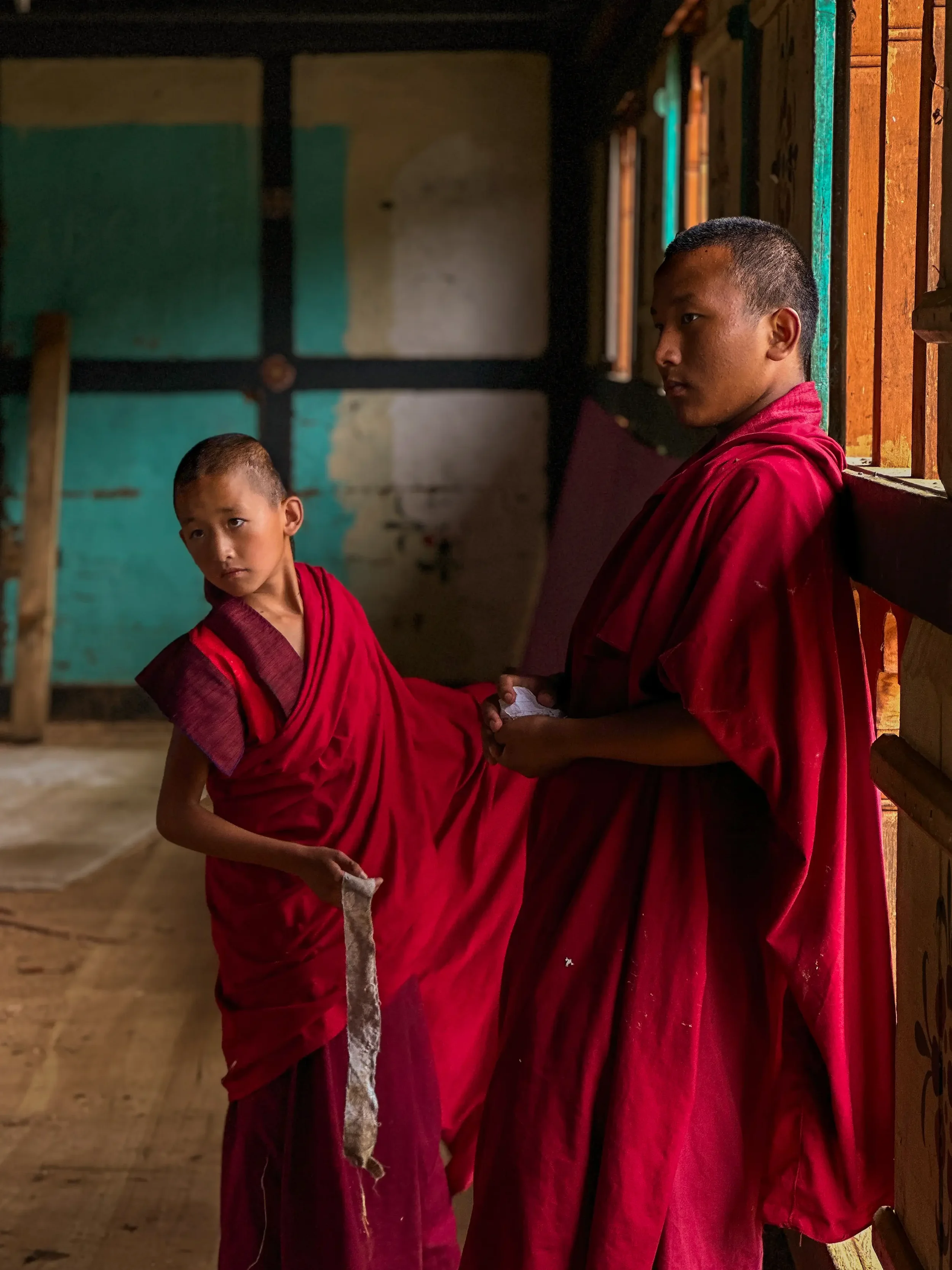Two young boys in red robes inside a wooden building, one is handing the other a cloth or paper. The boys have short hair and are standing near a window with wooden bars, with natural light illuminating the scene.