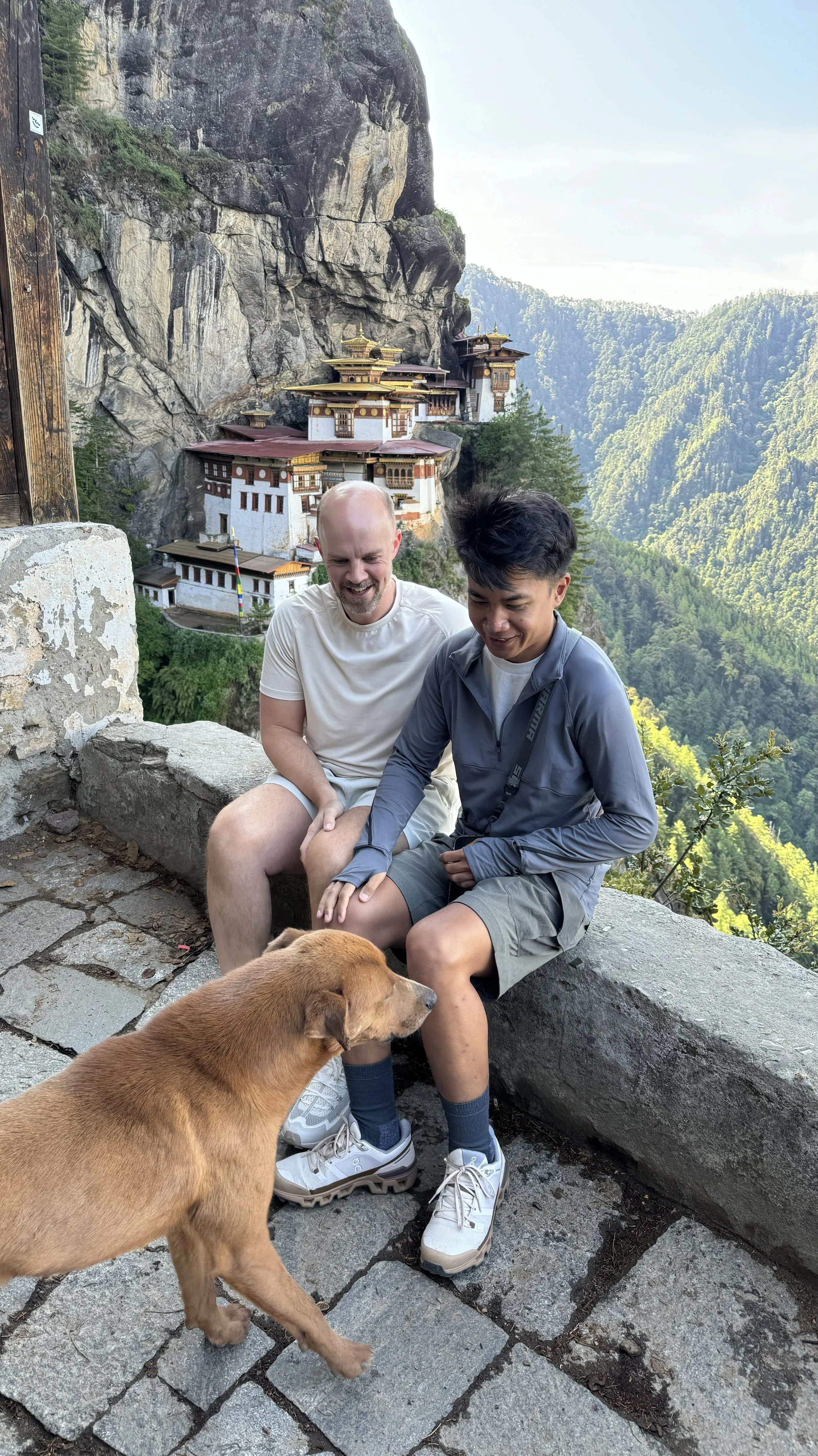 Matt and Wahyu sitting near Tiger’s Nest Monastery in Bhutan, with a dog in the foreground and forested mountains in the background.
