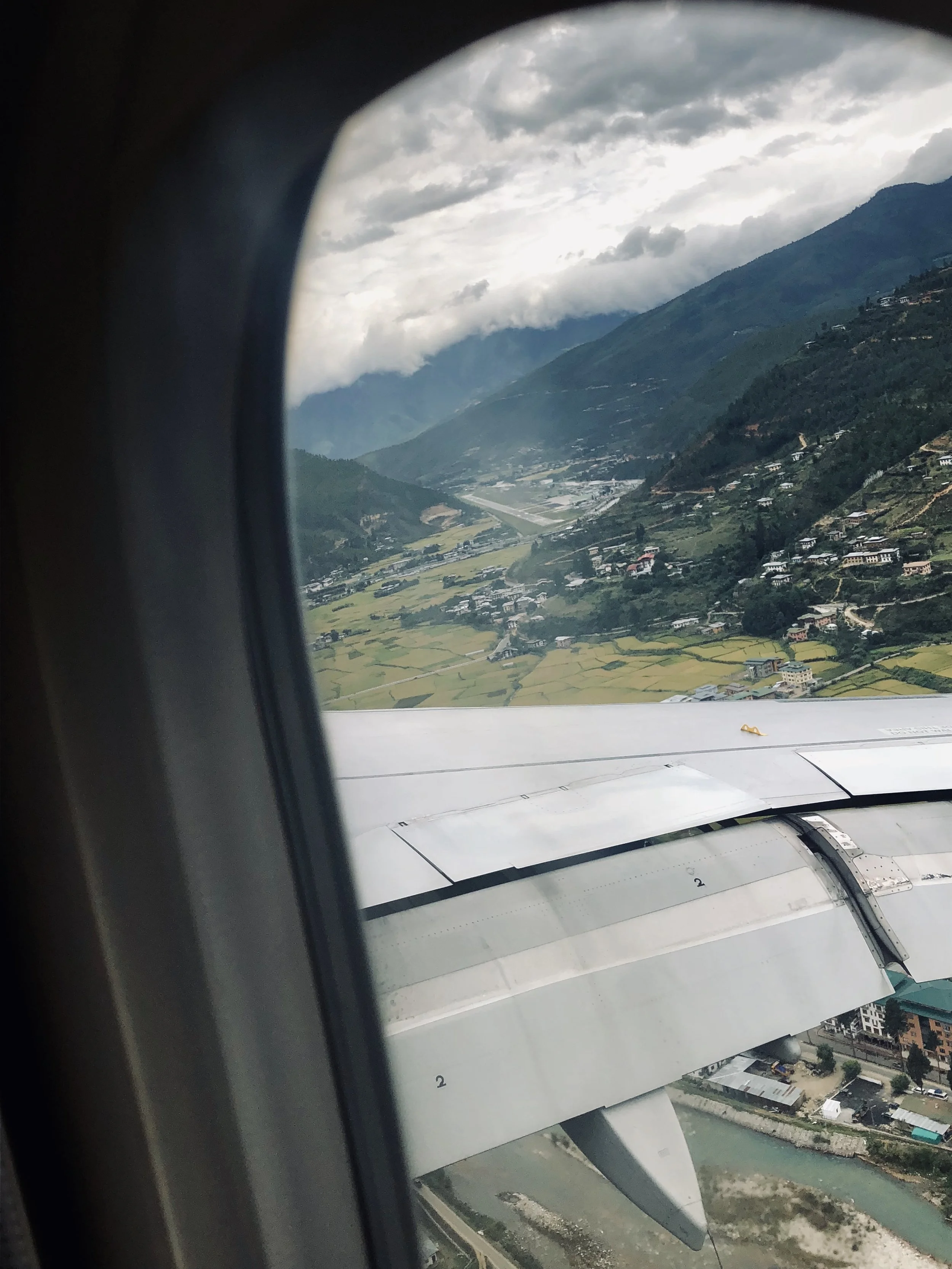 View from an airplane window while landing in Paro, Bhutan, with mountains, fields and the plane wing visible.