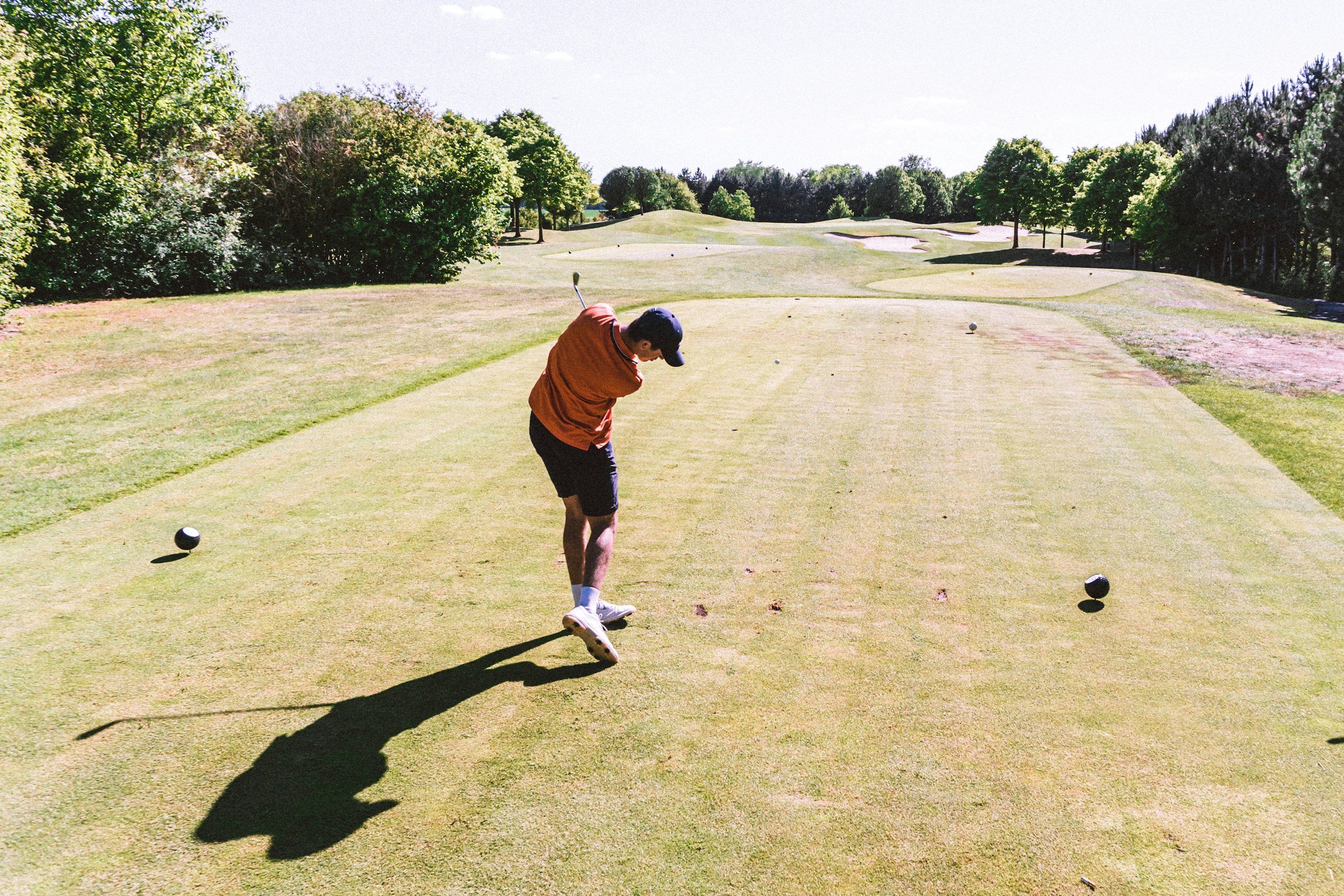 A man in a brown shirt, black shorts, and a black cap playing golf on a sunny golf course, preparing to hit a golf ball, with trees and a bright sky in the background.