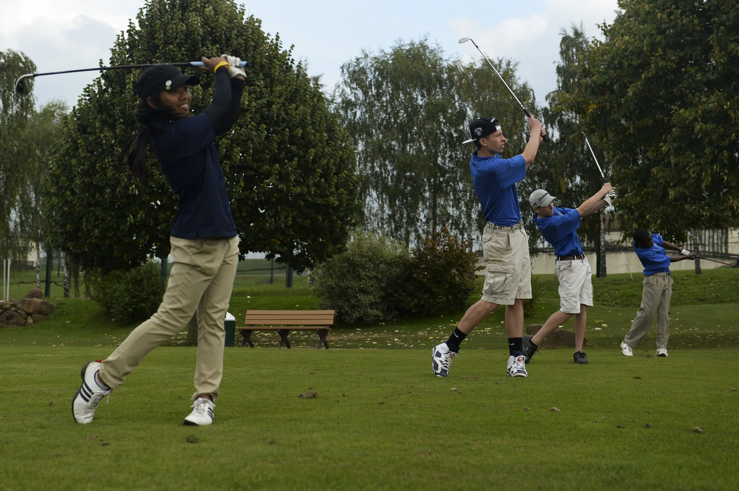 Four people practicing golf swings on a golf course, with trees and benches in the background.