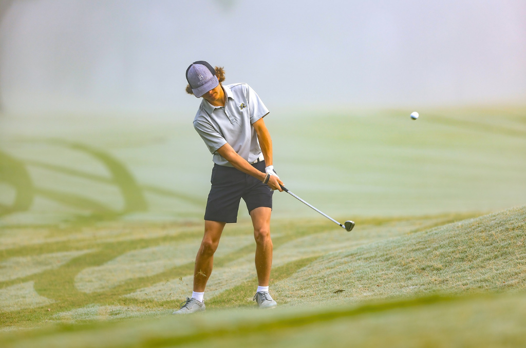 A woman playing golf on a course, swinging a club to hit a golf ball.
