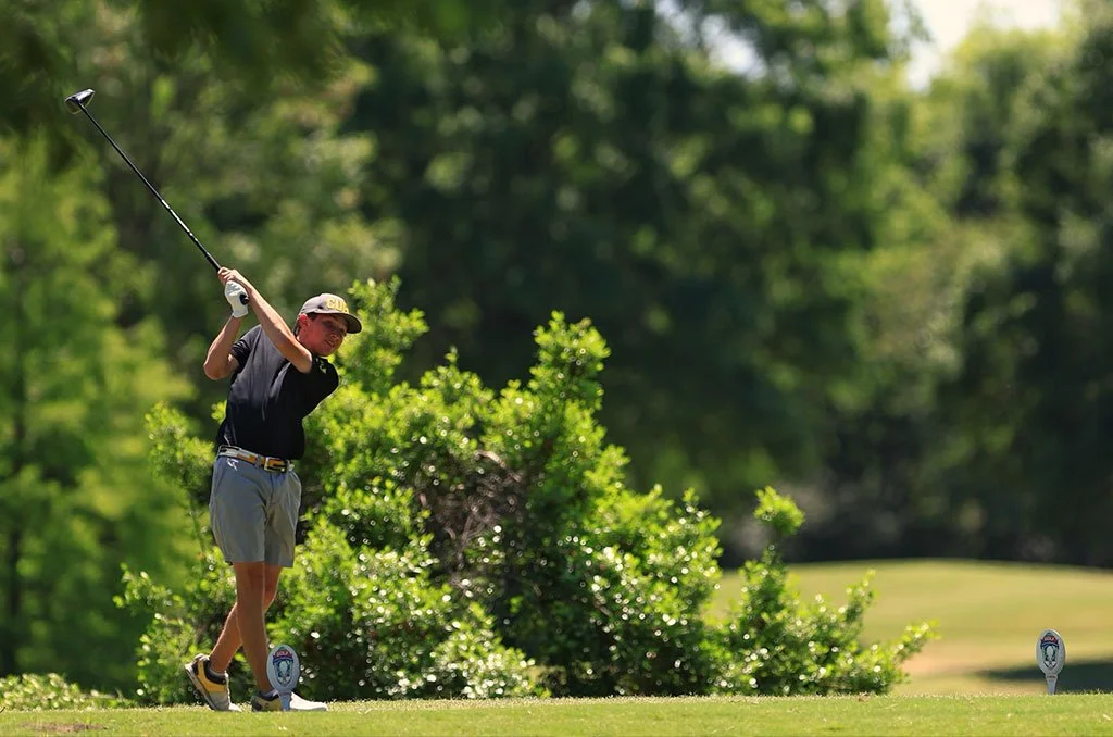 A person in golf attire is swinging a golf club on a golf course surrounded by greenery.
