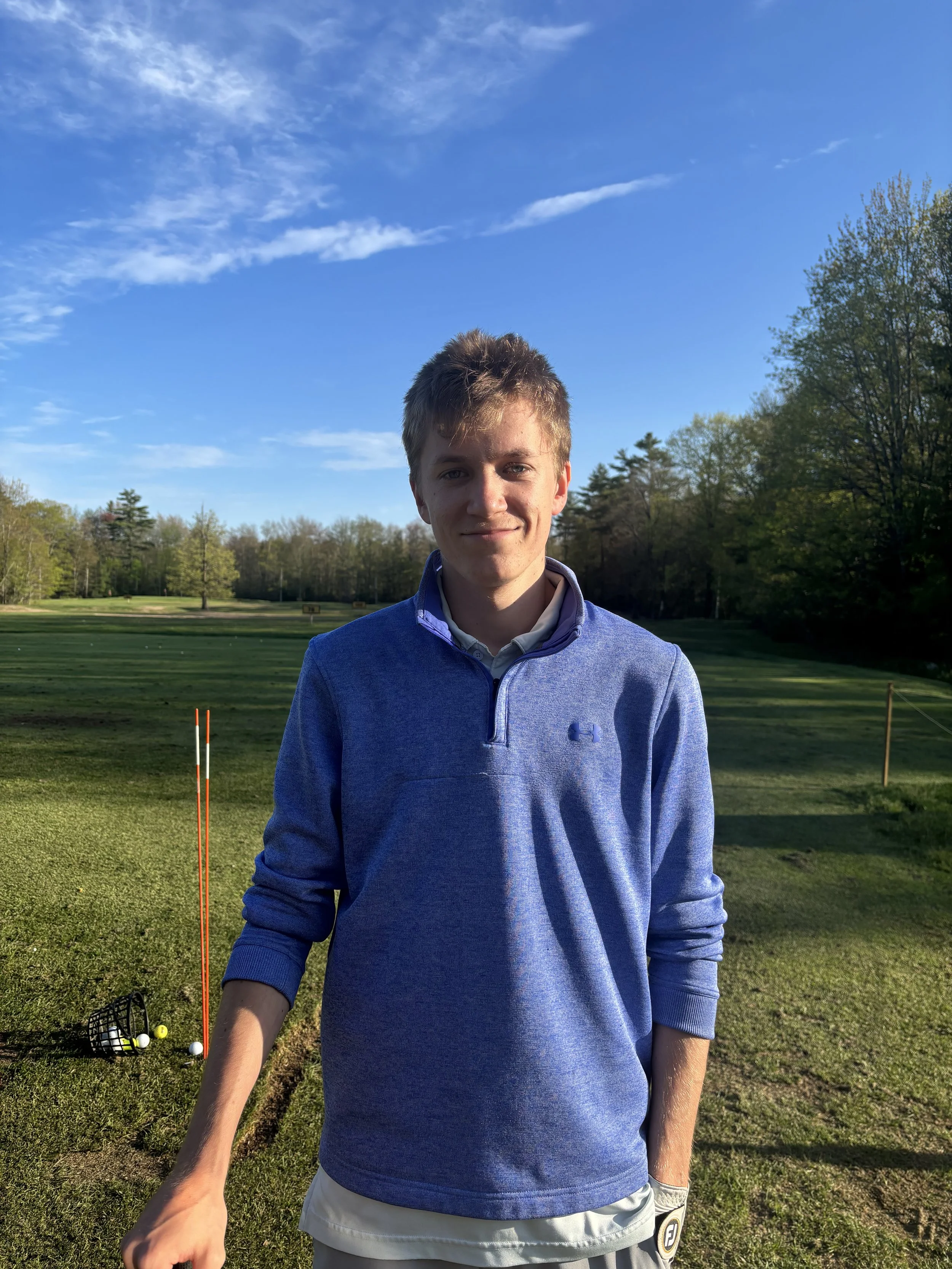 Young man in blue golf shirt standing on golf course with a basket of golf balls and golf flag in the background.