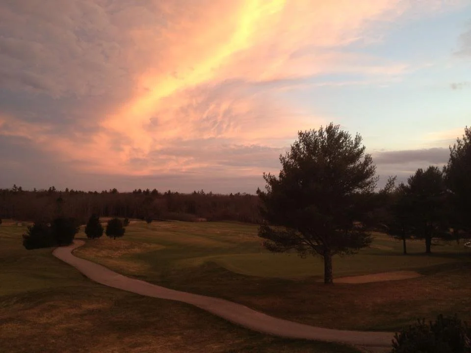 A scenic view of a golf course at sunset with a winding path, green grass, and trees under a colorful sky with pink and orange clouds.
