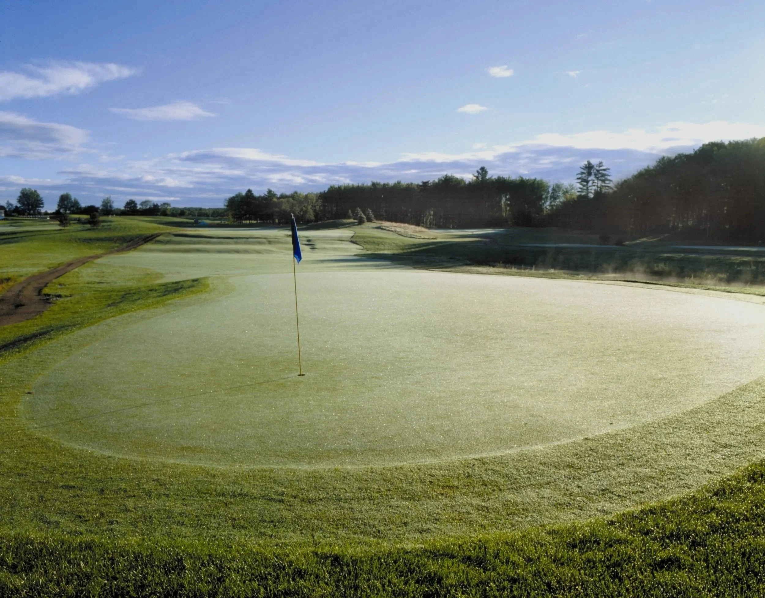 A golf course green with a flag, surrounded by fairways and trees under a blue sky with clouds.