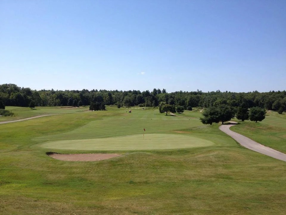A view of a golf course with a putting green, sand trap, fairway, and trees under a clear blue sky.
