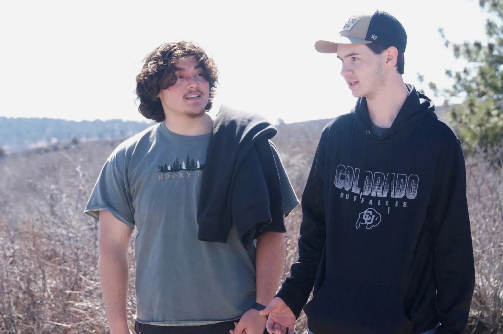 Two young men walking outdoors on a sunny day, holding hands, with trees and a hill in the background.