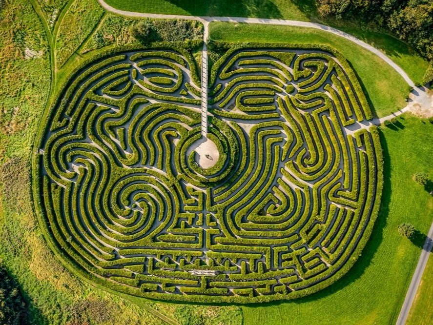 Aerial view of a heart-shaped maze in a park with green hedges and a circular central area connected to a pathway.