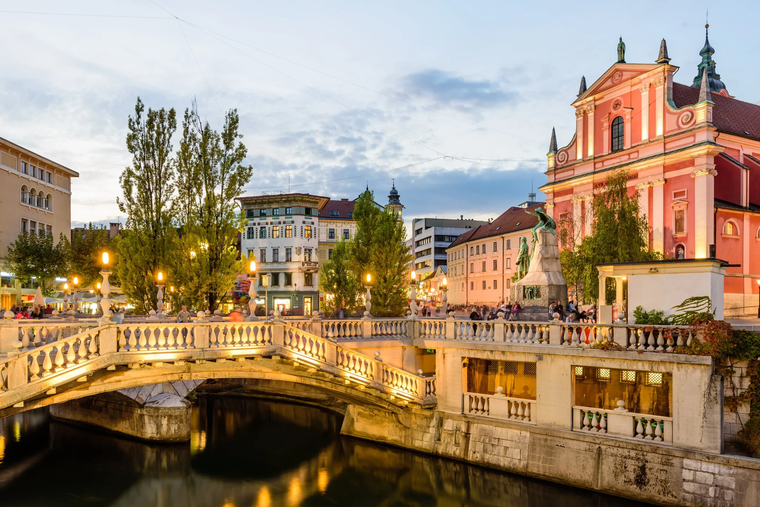 A city square at dusk featuring a pink Baroque church with statues and ornate architecture, a small bridge crossing a canal, and surrounding buildings with people walking and sitting under decorative street lamps.