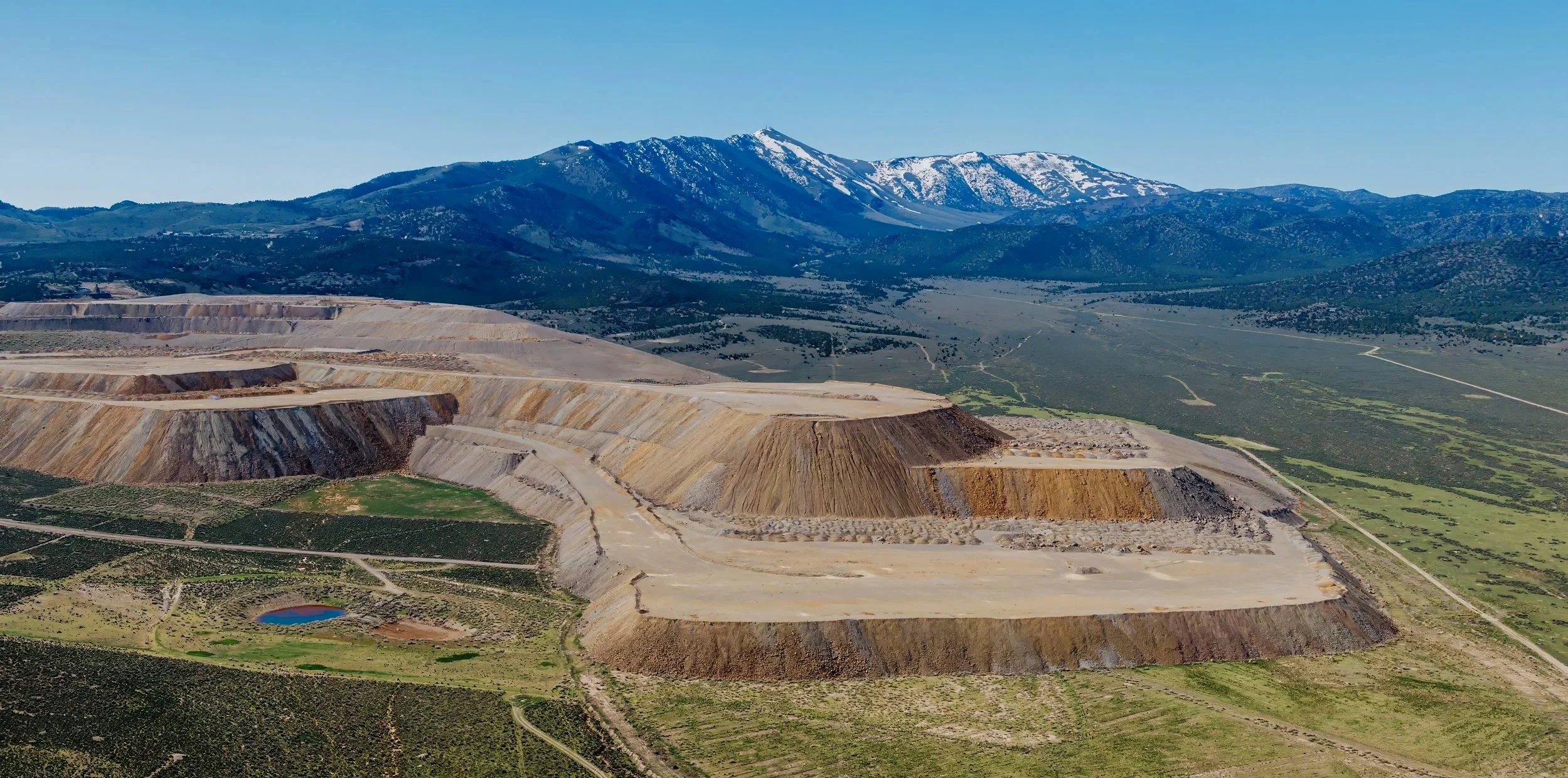 A large open pit mining operation with terraced levels, surrounded by green fields, with a mountain range with snow-capped peaks in the background under a clear blue sky.