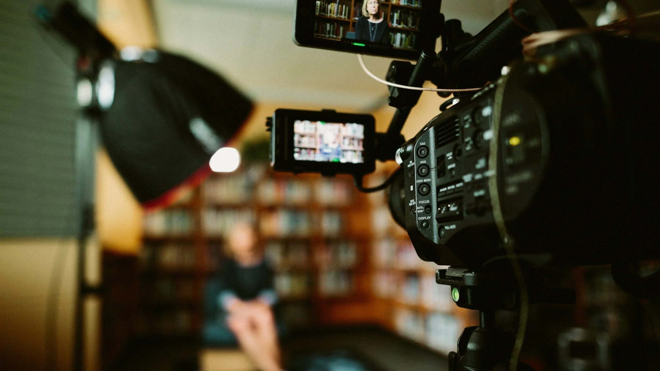 A professional video camera filming a woman giving a presentation or interview in a library filled with books.