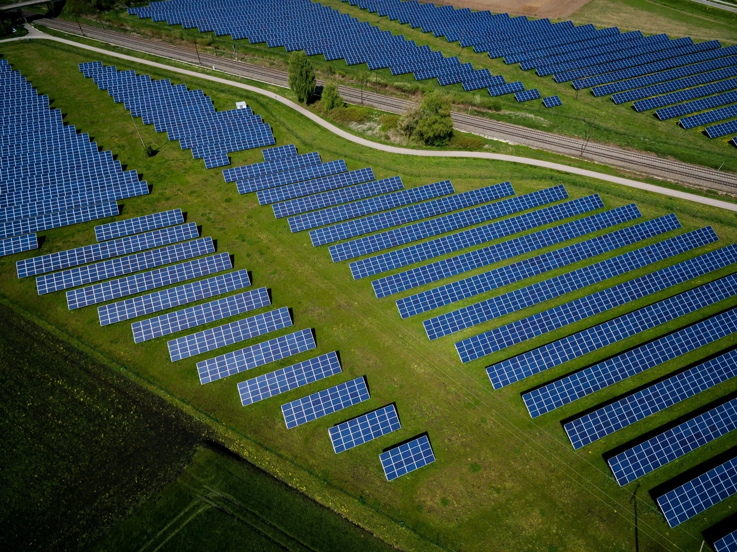 An aerial view of a solar farm with numerous blue solar panels arranged in rows on grassy land, with a path and railway tracks running through the middle.