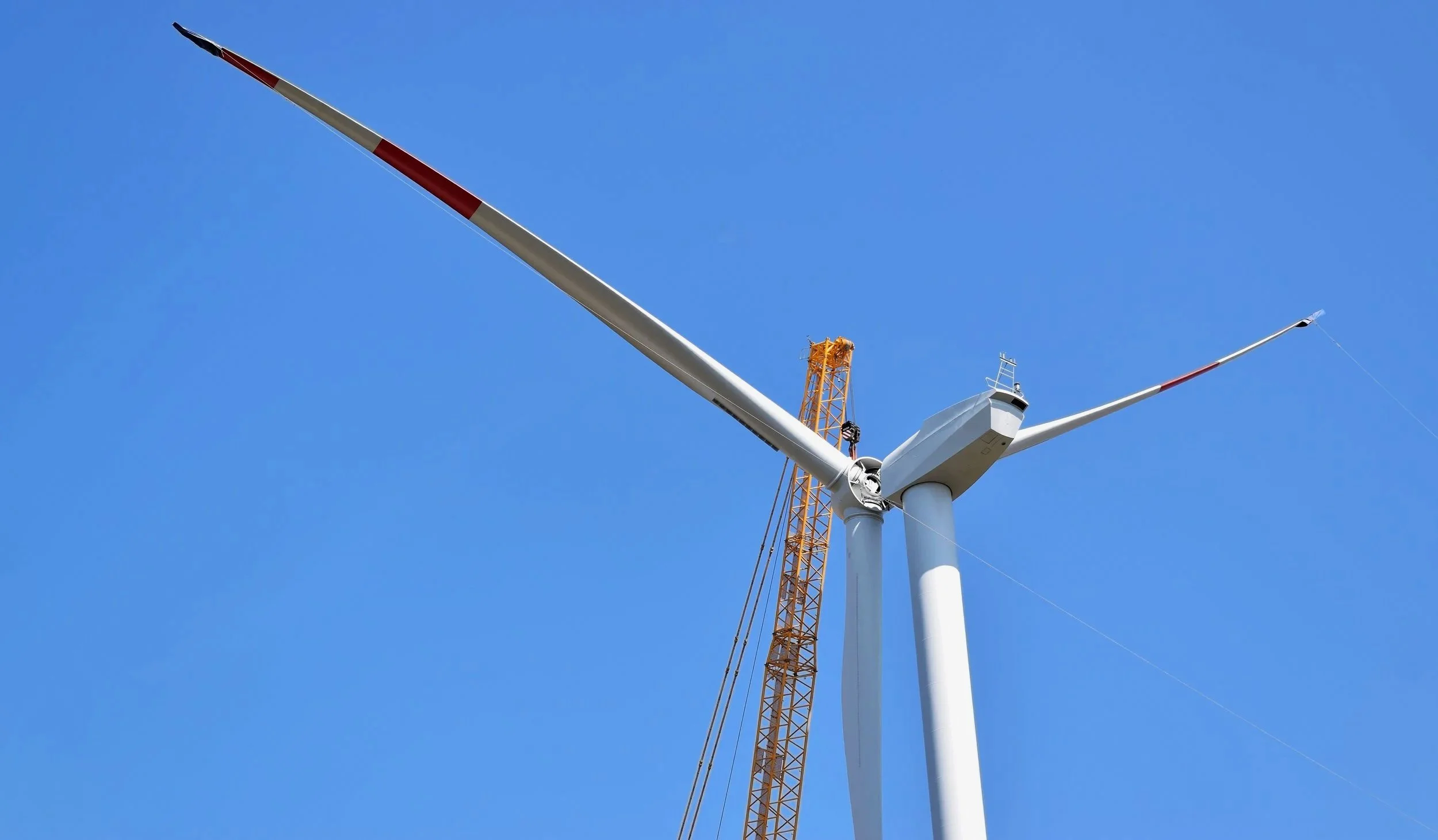 Wind turbine under construction with a yellow crane in the background and a clear blue sky.