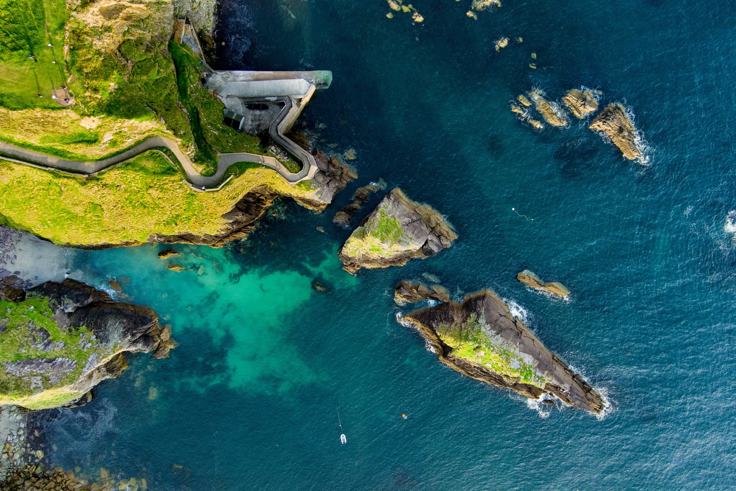 An aerial view of a coastal area with a narrow winding road, green grassy cliffs, a concrete structure, and rocks in the ocean. There are a few boats in the water.