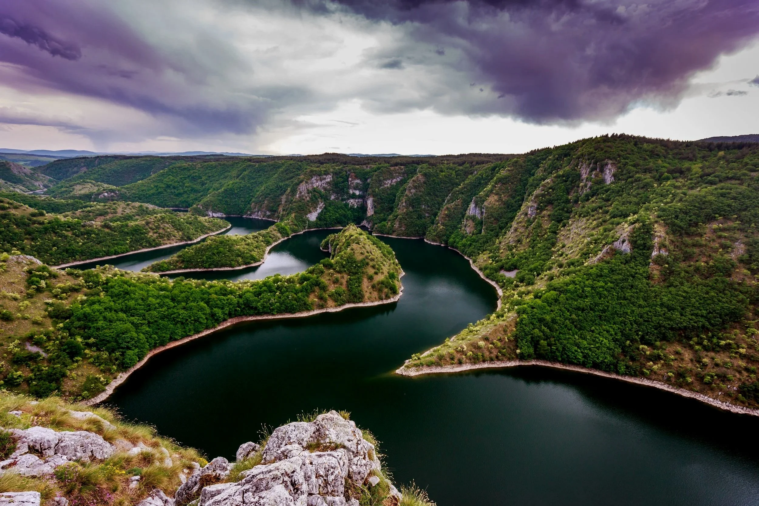 A winding river flows through green rolling hills under a dark, stormy sky.