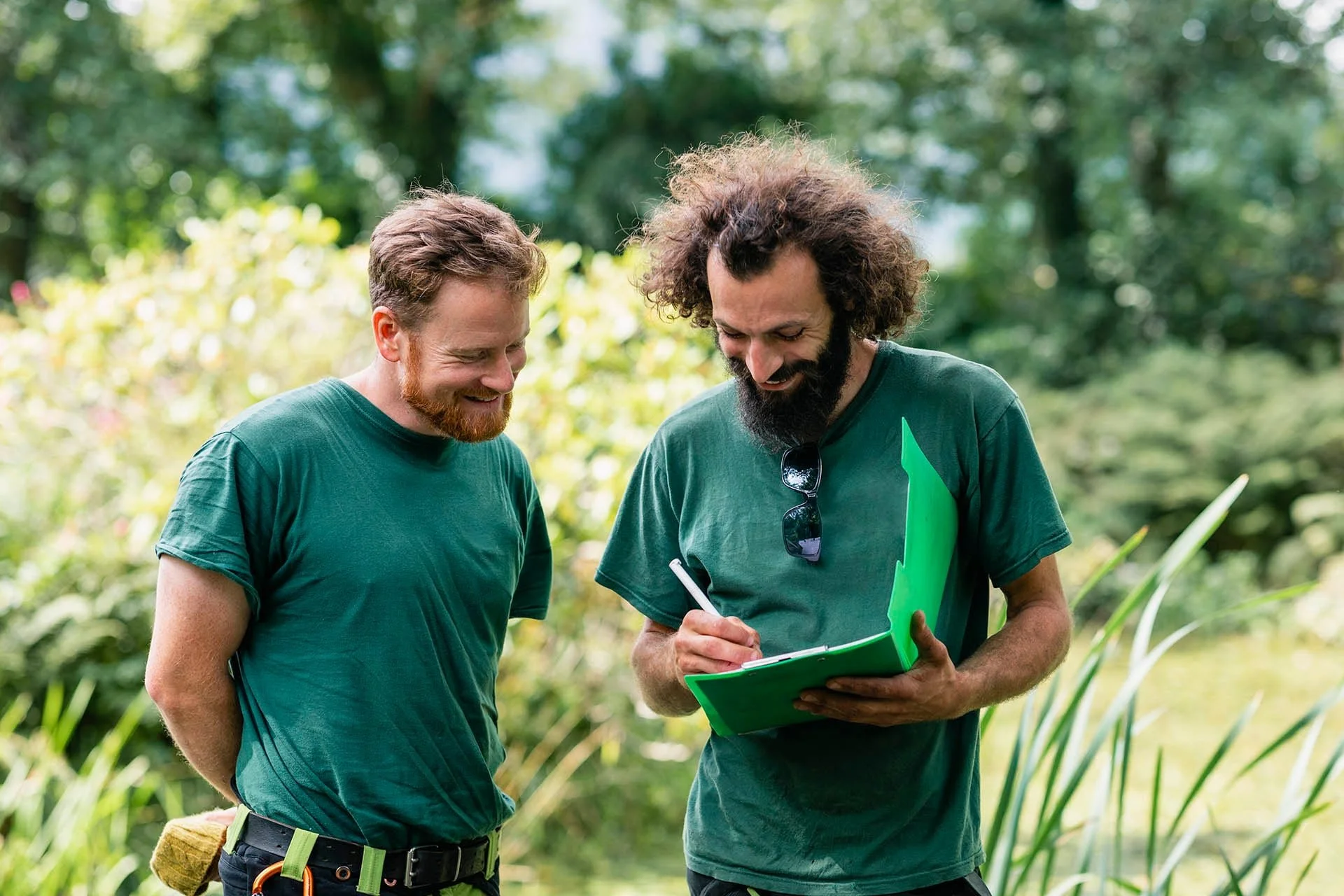 Two men with beards and curly hair standing in a lush green outdoor setting, looking at a clipboard held by one of them. Both are wearing green shirts, one has sunglasses hanging from his shirt, and the other has a yellow towel tucked into his waistband. They appear to be discussing something related to gardening or outdoor activity.