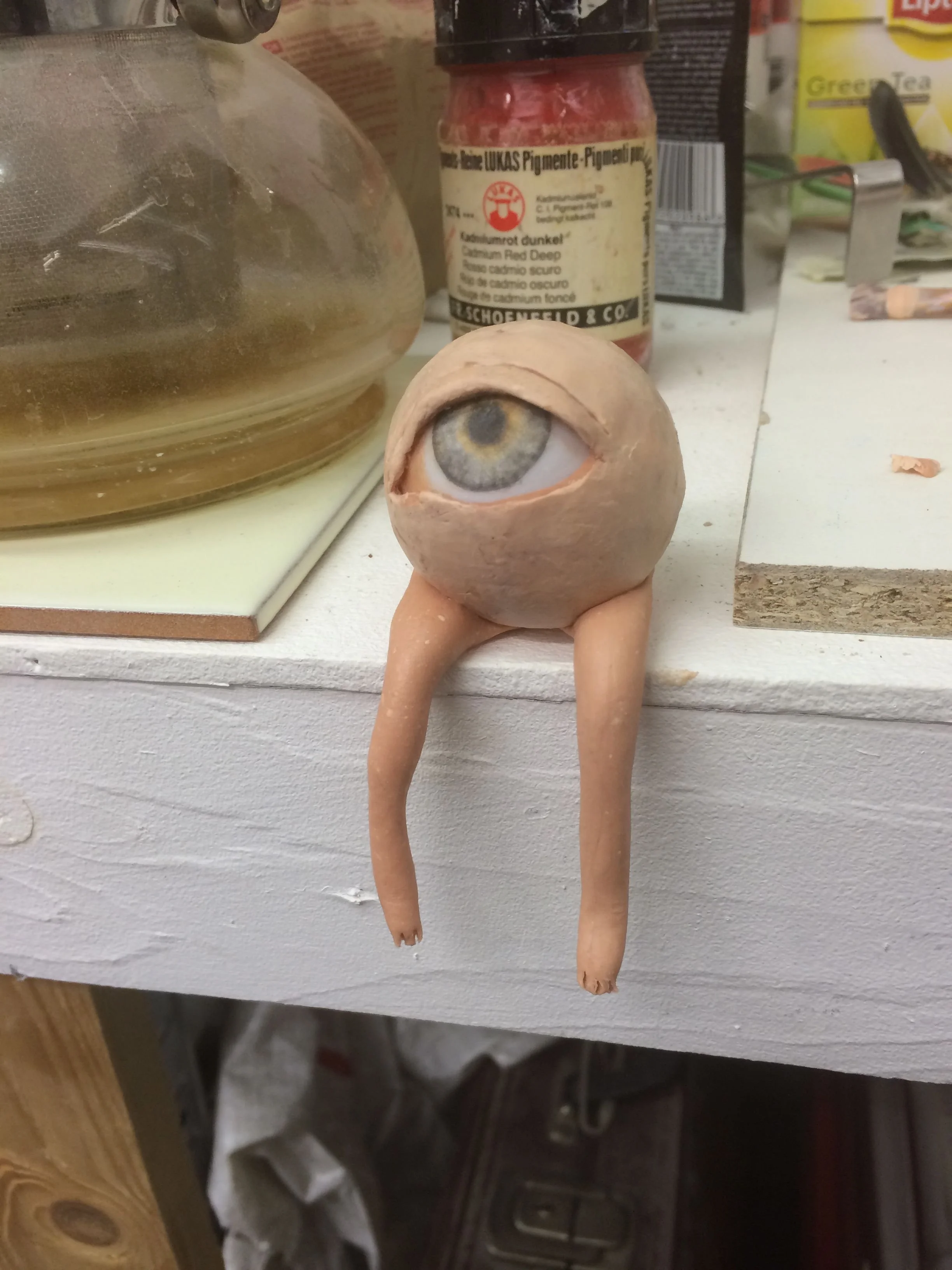 A small sculpture with a large realistic human eye on top and two small clay legs hanging down, sitting on a white wooden surface surrounded by containers and miscellaneous items.