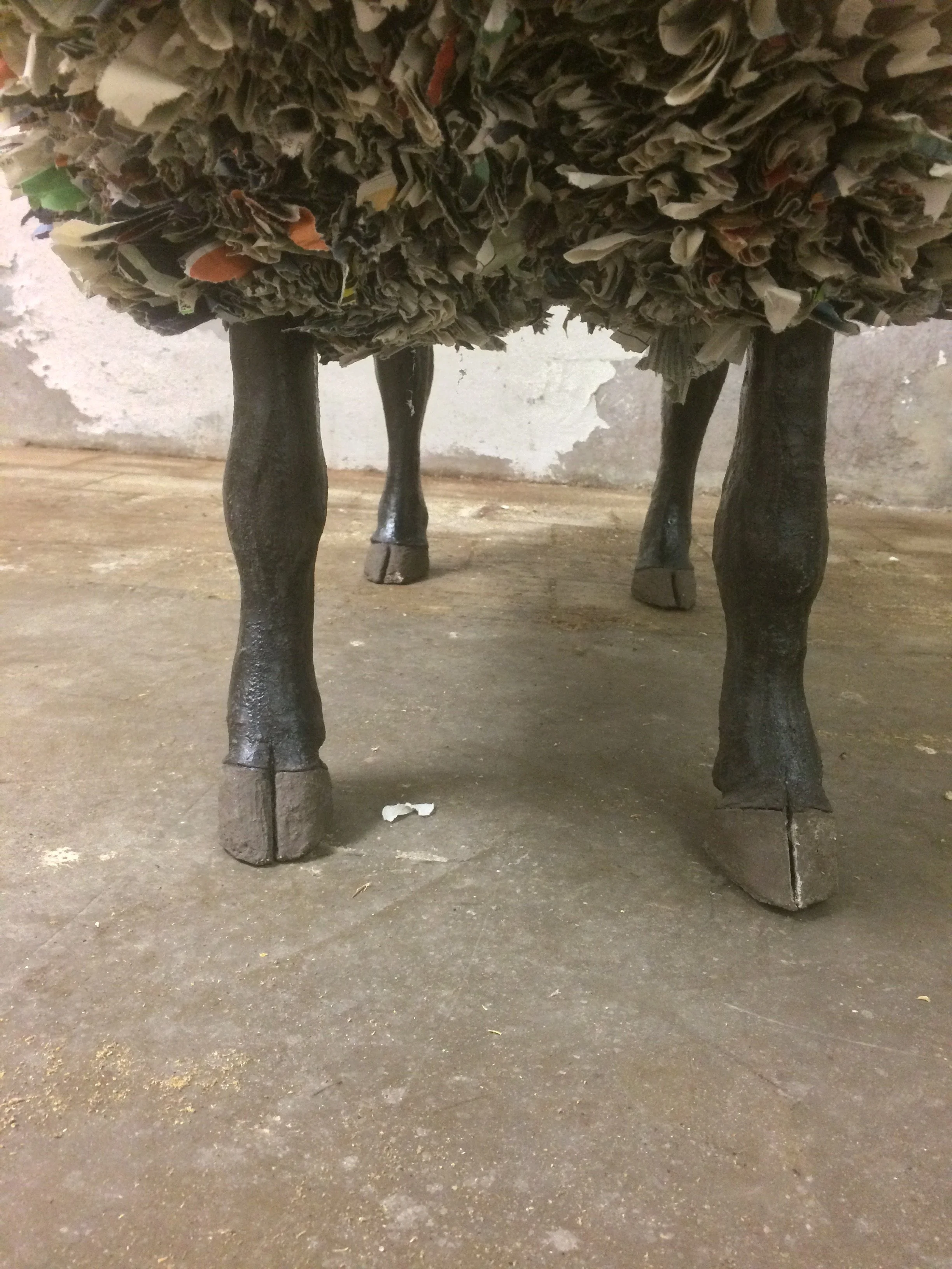 Close-up of a table with four wooden legs, with the top covered in crumpled paper or fabric, against a worn wall and cement floor.