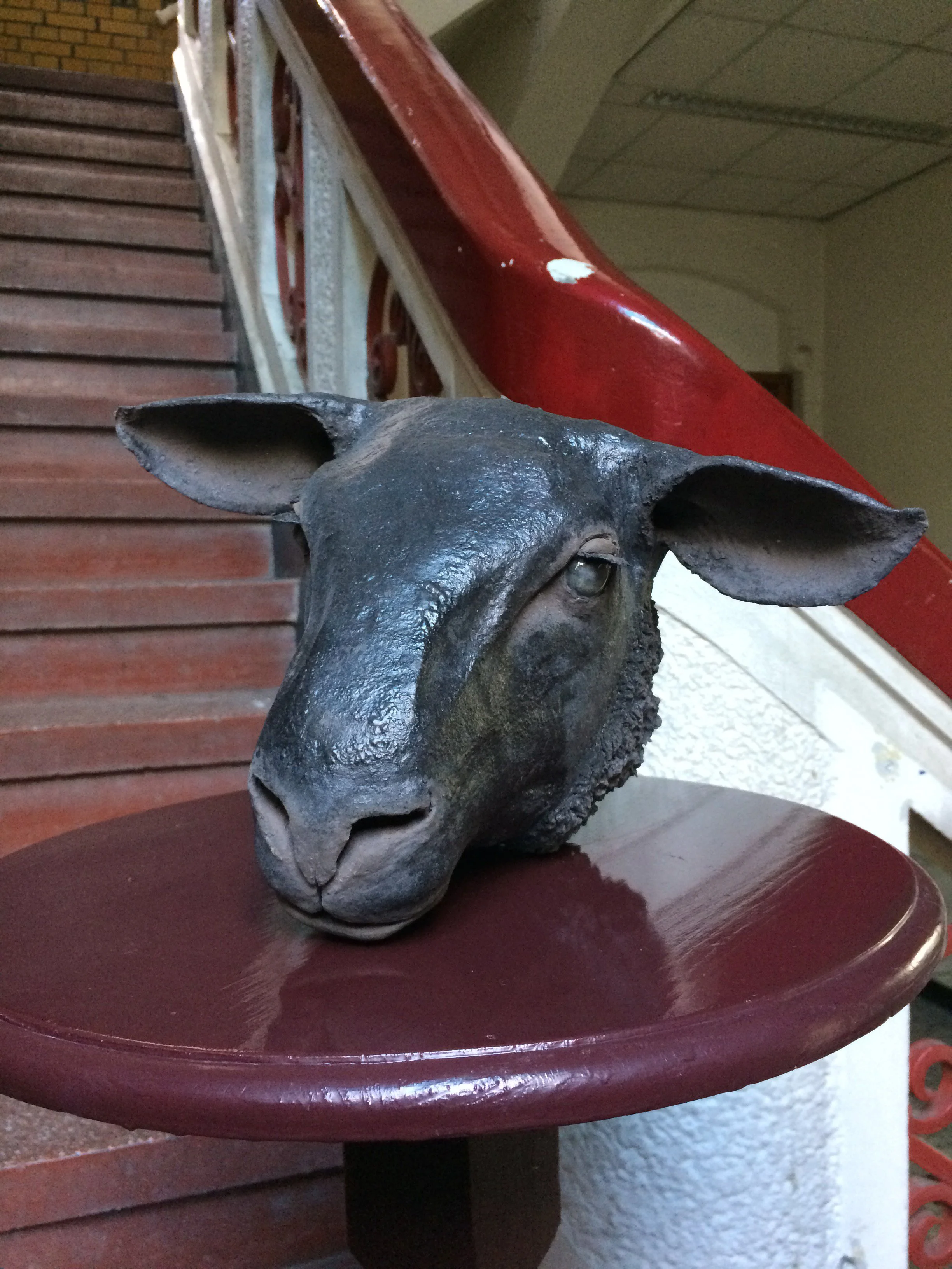 Photograph of a sculpture of a horse's head placed on a small round table, with a background featuring a staircase with red handrails and wooden steps.